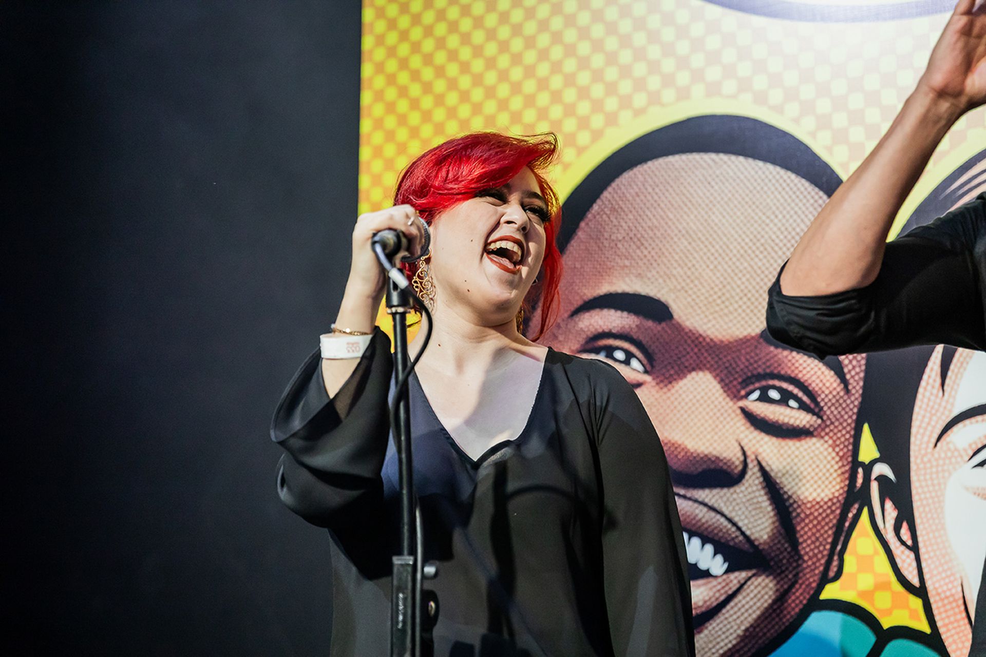 Uma artista com cabelo vermelho vivo cantando em um microfone no palco, em frente a um cenário com estilo de quadrinhos.