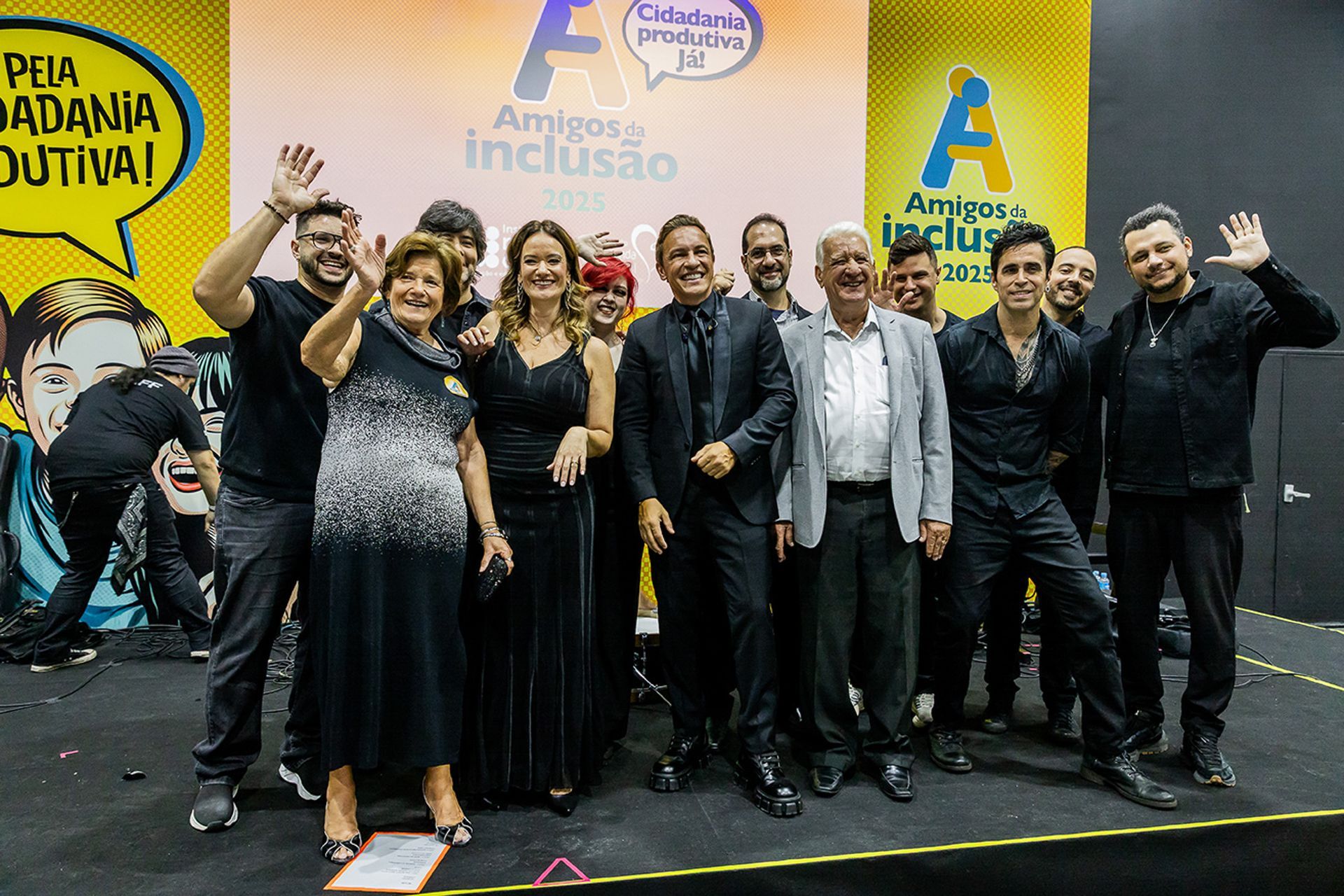 Um grupo de pessoas em pé no palco durante um evento da Amigos da Inclusão, sorrindo e acenando para o público.