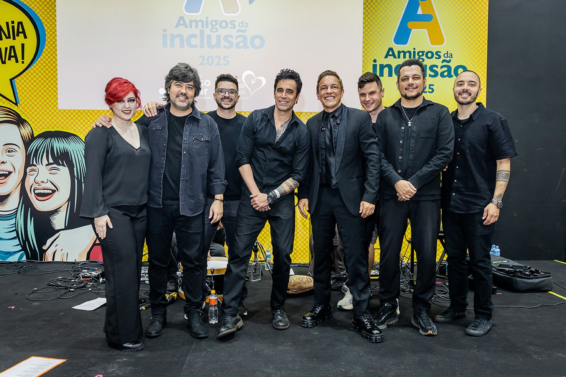 Oito pessoas posam para uma foto de grupo em um palco em frente a uma faixa amarela para um evento da Amigos da Inclusão.