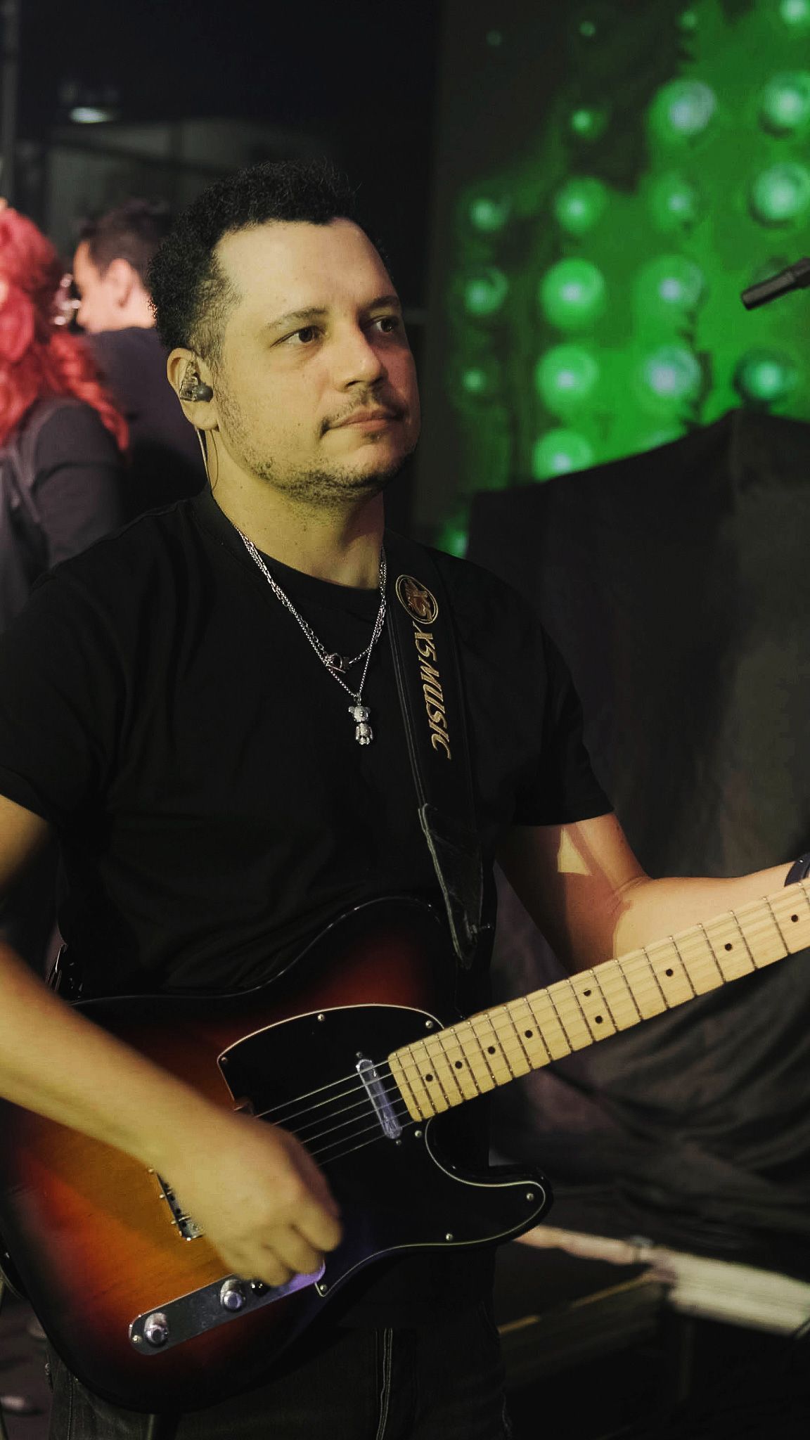Uma pessoa tocando uma guitarra elétrica sunburst enquanto se apresenta no palco com iluminação verde ao fundo.