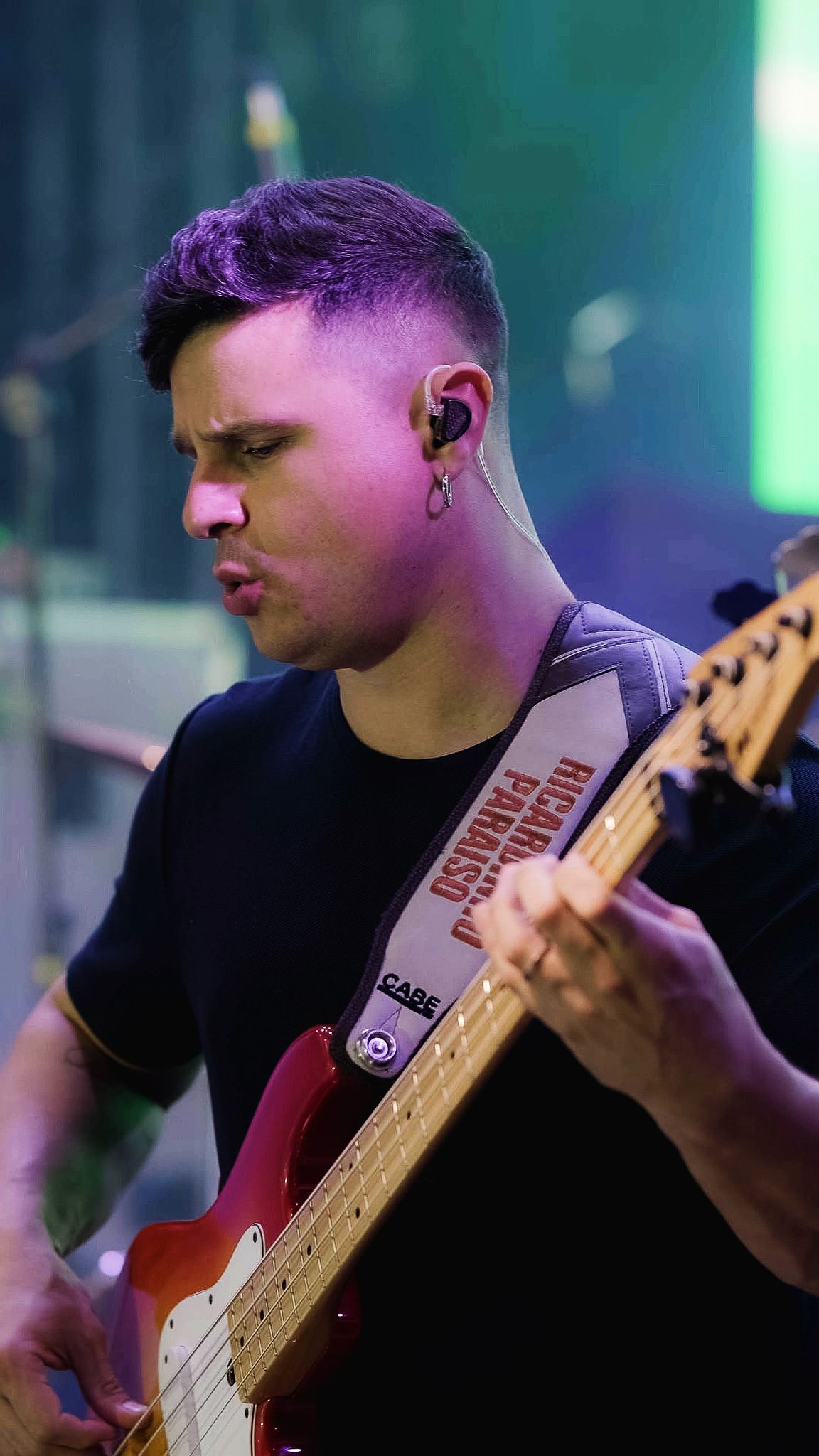 Um músico toca um baixo elétrico vermelho no palco, vestindo uma camisa preta e um fone de ouvido intra-auricular.