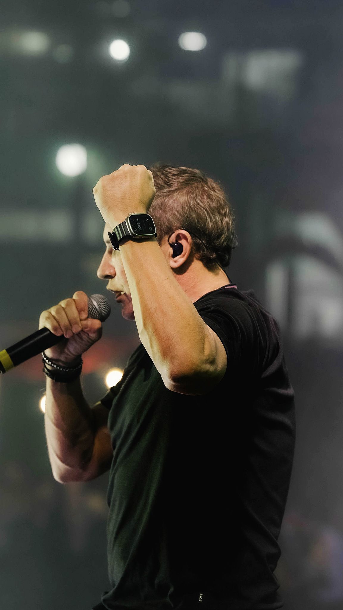 Uma pessoa vestindo uma camisa preta cantando em um microfone, com um punho erguido no ar durante um show.