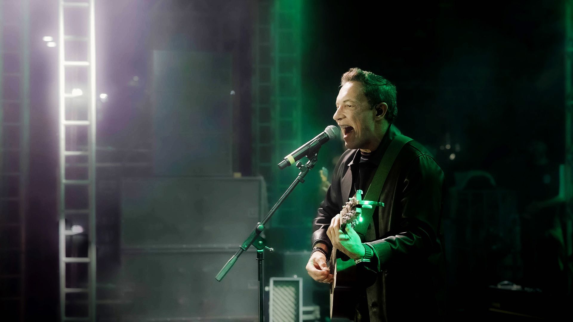 Um músico cantando ao microfone enquanto toca violão em um palco com iluminação verde e tênue.