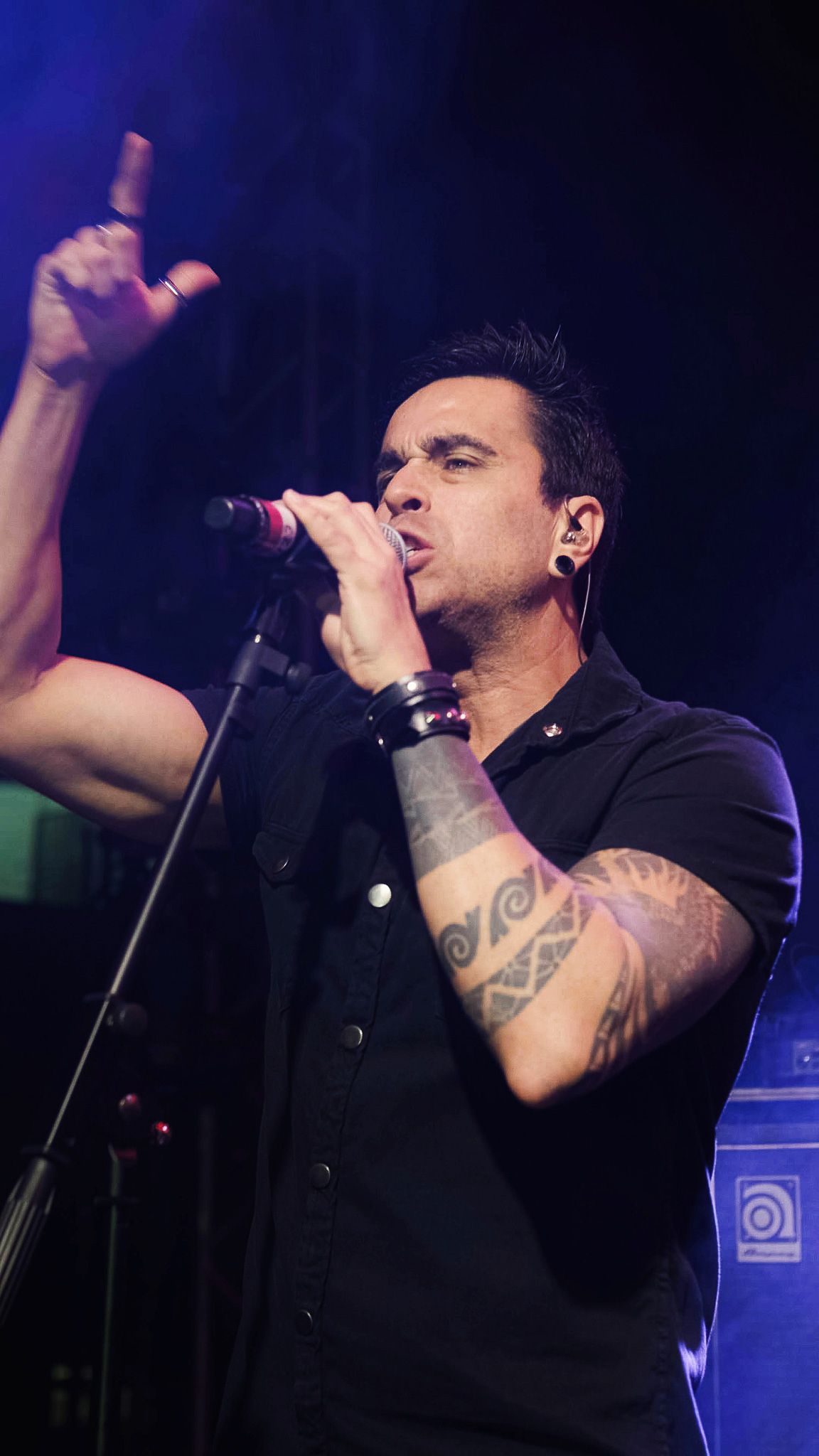 Um cantor se apresenta no palco, segurando um microfone, apontando para cima com uma das mãos e exibindo tatuagens.