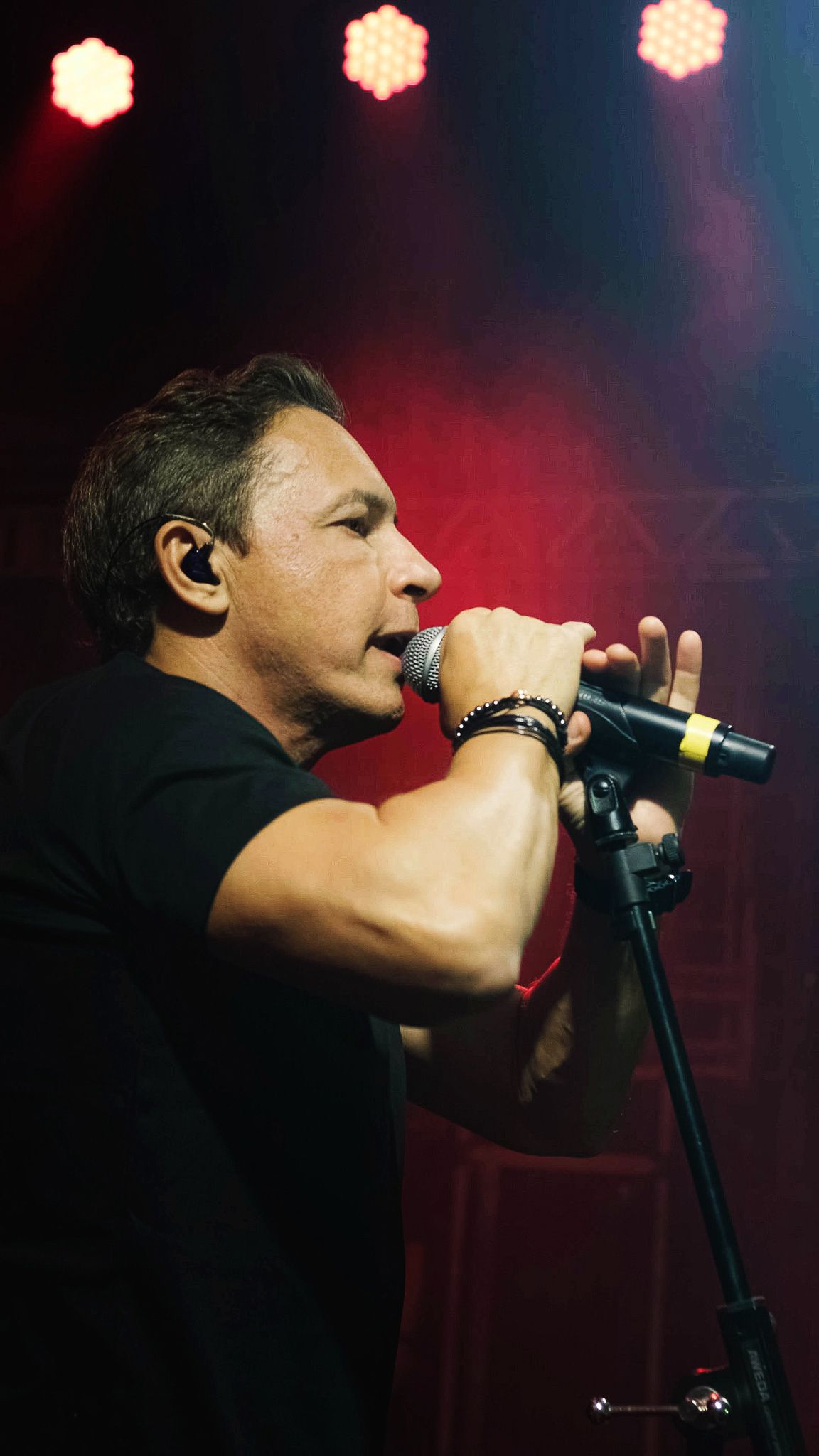 Um músico de camisa preta canta em um microfone no palco sob holofotes vermelhos brilhantes.