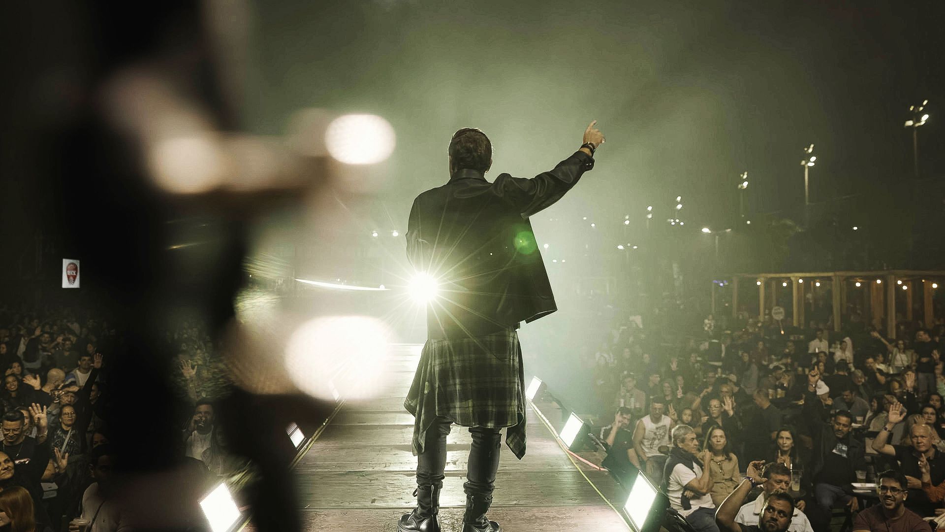 Um artista no palco, visto de costas, levanta uma das mãos para uma grande plateia sob luzes de palco brilhantes e intensas.