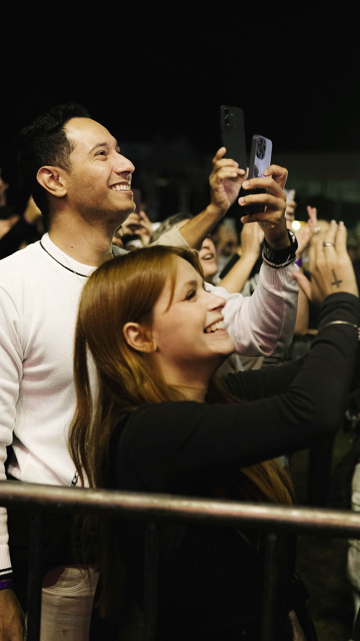 Duas pessoas sorrindo enquanto seguram seus celulares para registrar um evento em um ambiente com pouca luz.