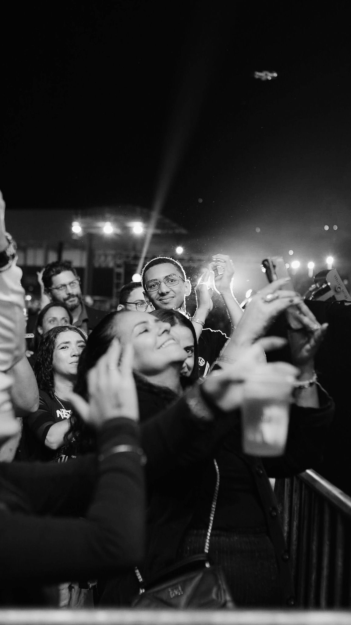 Uma imagem em preto e branco de uma multidão em um show, com pessoas vibrando, aplaudindo e gravando com smartphones.