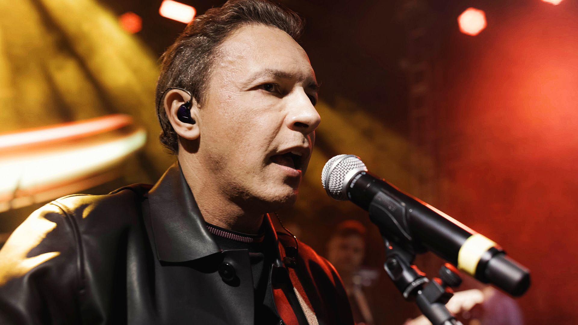 Um músico canta ao microfone no palco, vestindo uma jaqueta de couro preta sob luzes vermelhas.