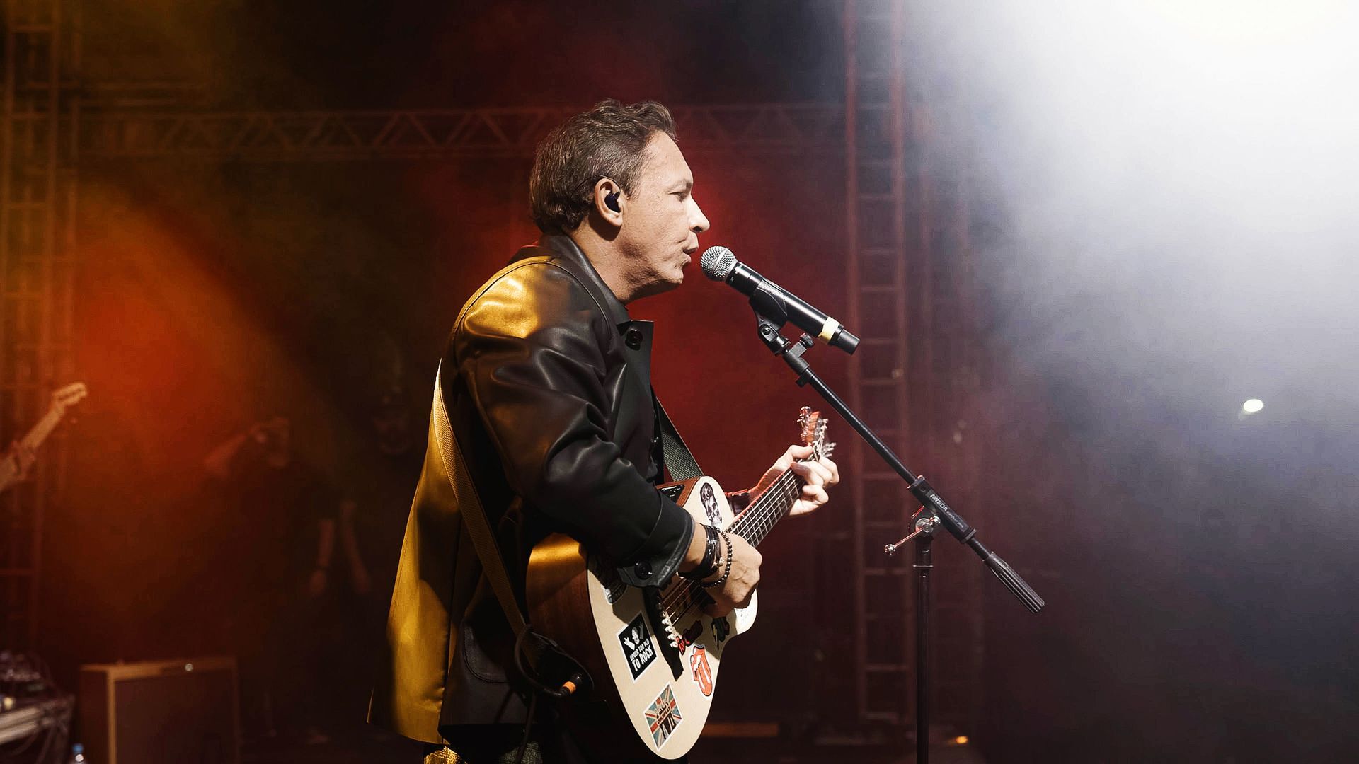 Um músico de jaqueta de couro toca violão e canta ao microfone em um palco com iluminação dramática.