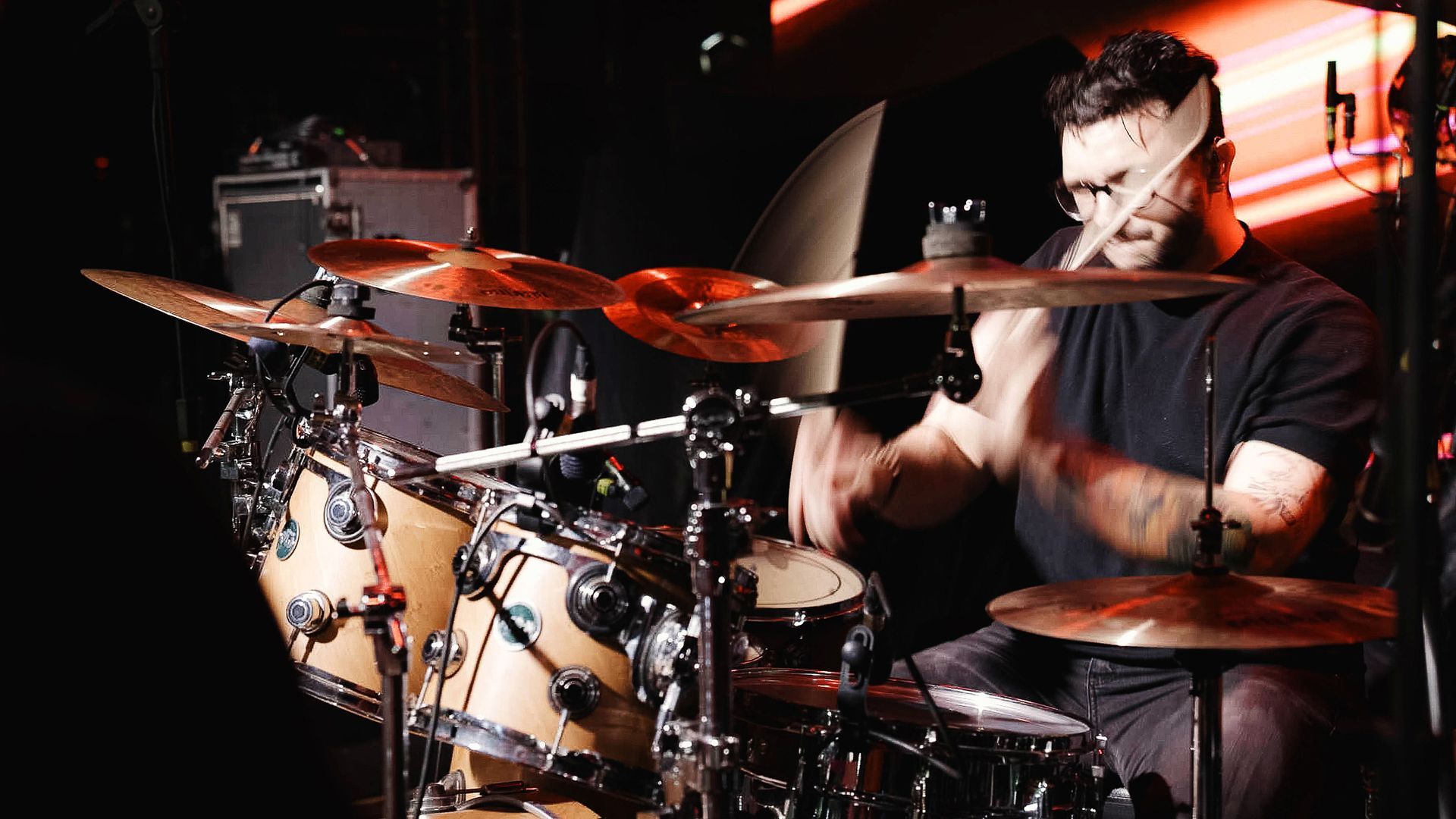 Um baterista concentrado toca uma bateria de cor clara com pratos de bronze em um palco profissional com pouca luz.