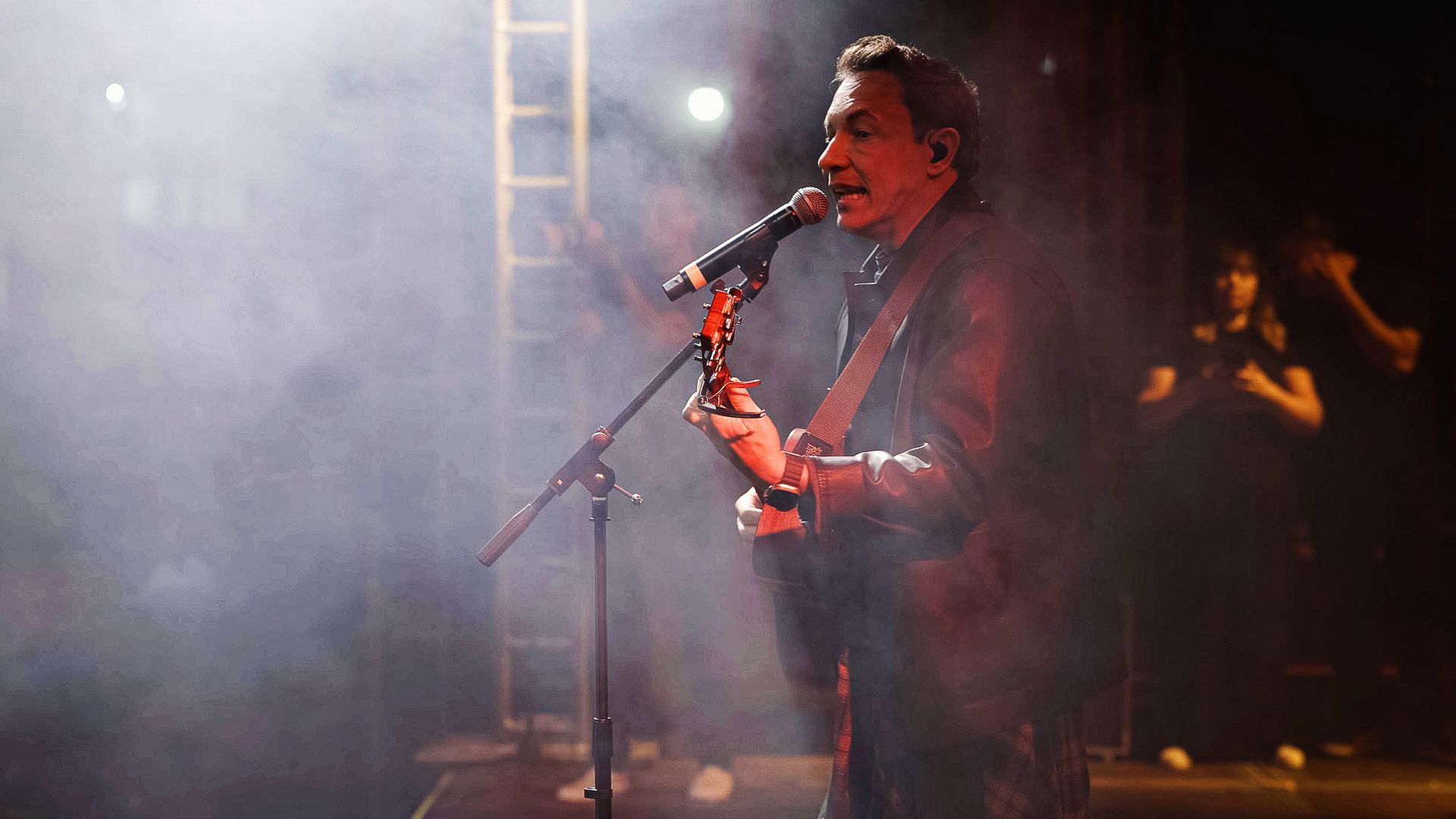Um músico toca violão e canta em um microfone em um palco pouco iluminado e esfumaçado.