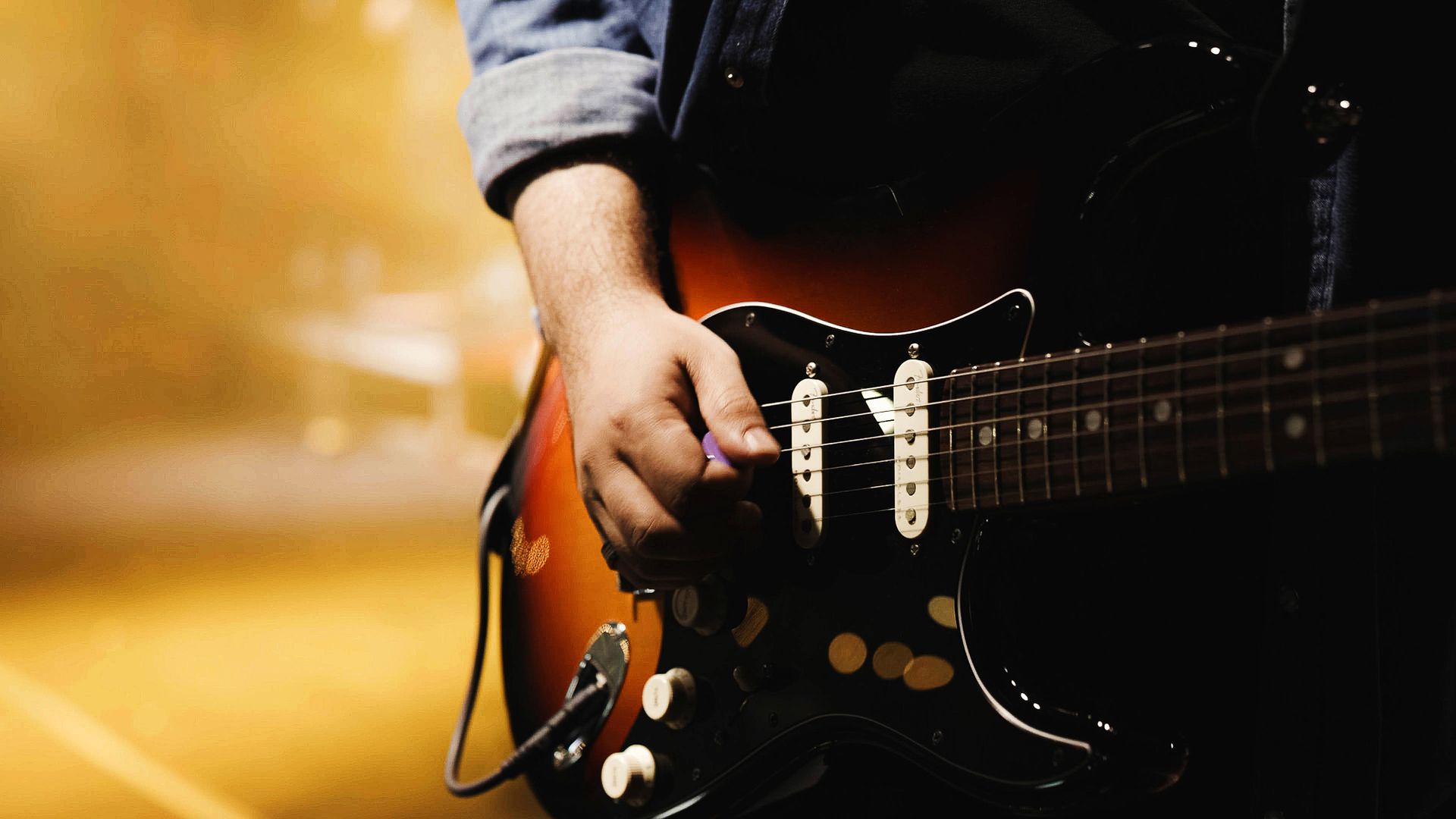 Primeiro plano de uma pessoa tocando uma guitarra elétrica sunburst contra um fundo quente e desfocado.