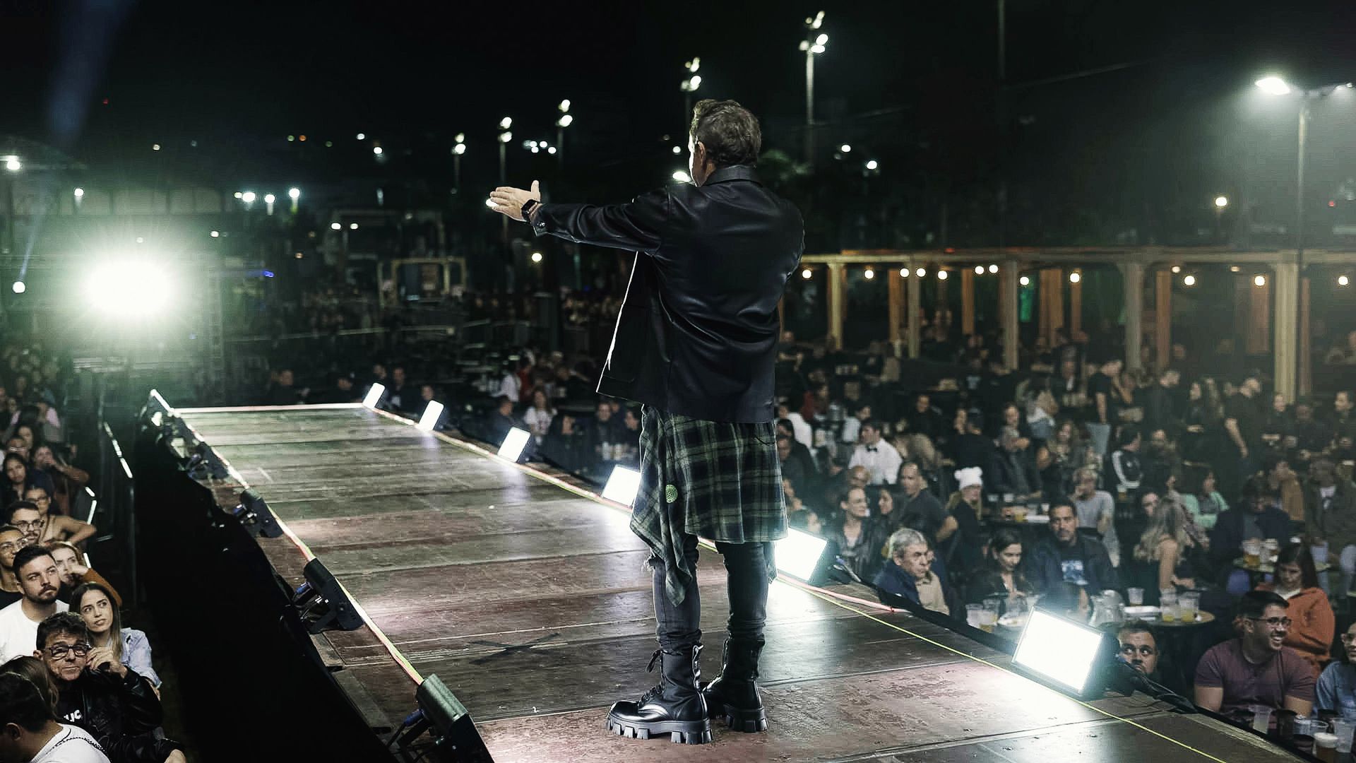Artista em um palco com os braços estendidos, de frente para uma plateia sentada em um local externo com pouca luz à noite.