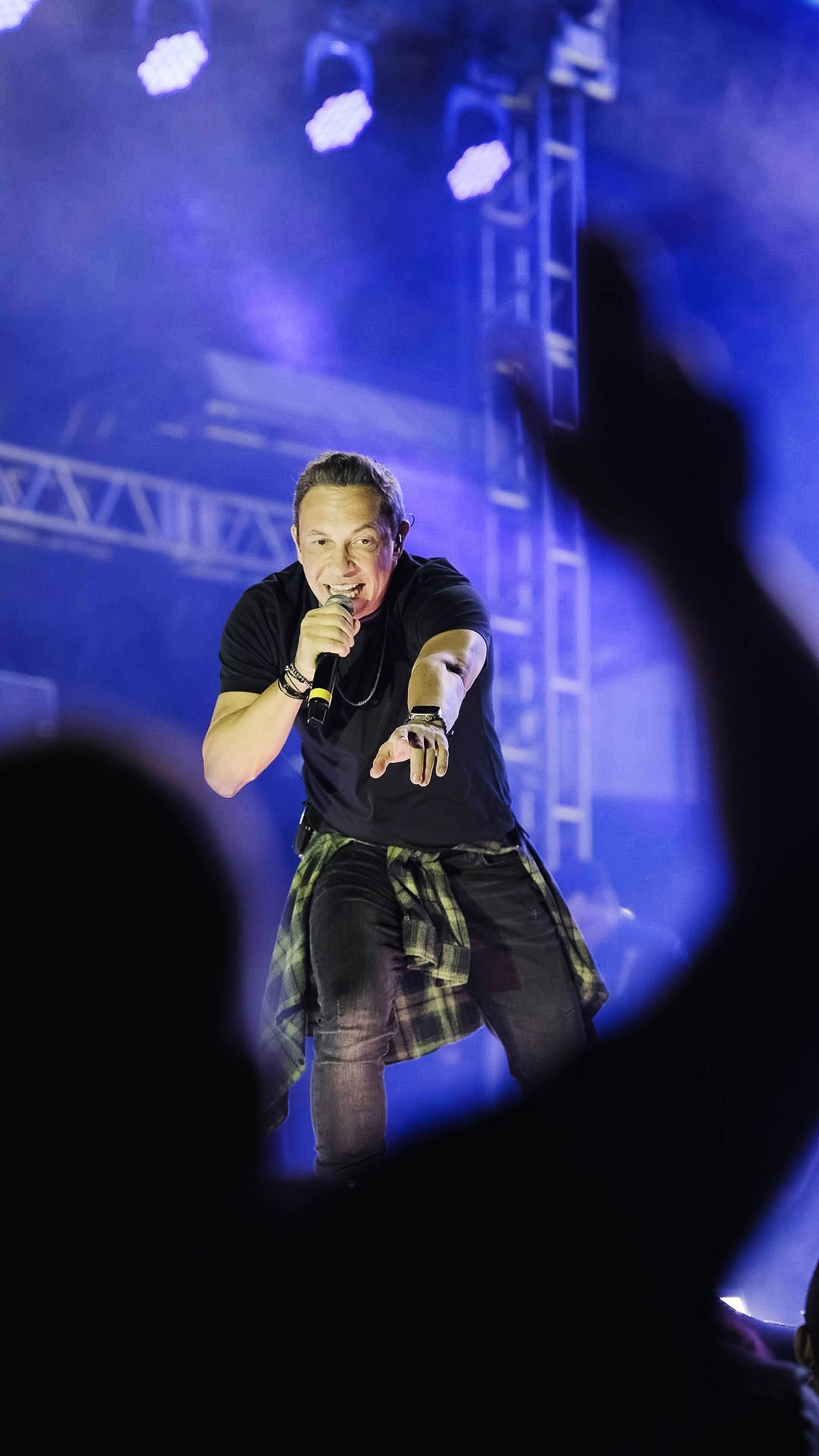 Uma artista canta em um microfone em um palco iluminado com luzes azuis, com a silhueta desfocada de um fã em primeiro plano.