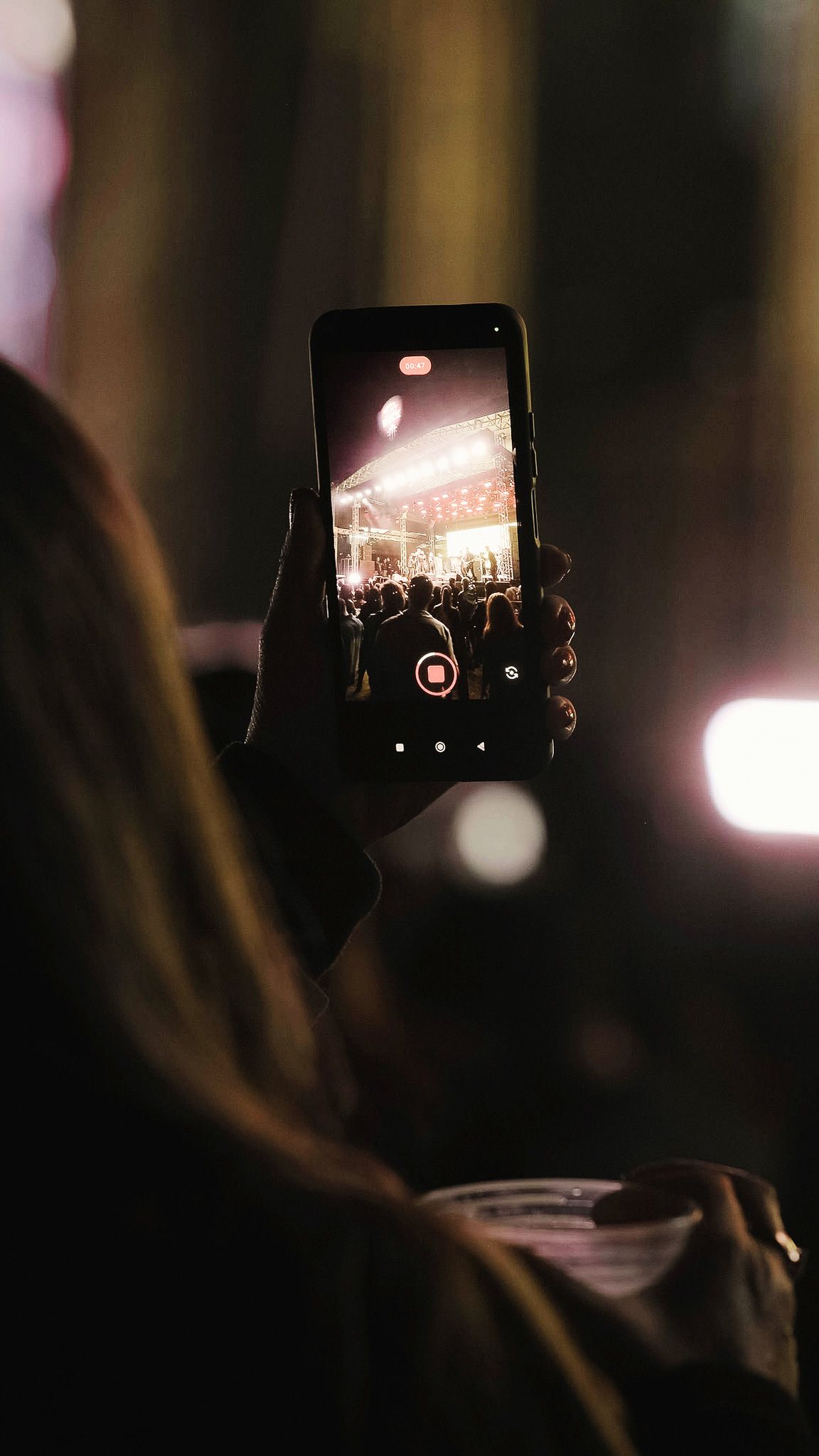 Uma pessoa segurando um celular para gravar um palco de show com luzes fortes à noite.