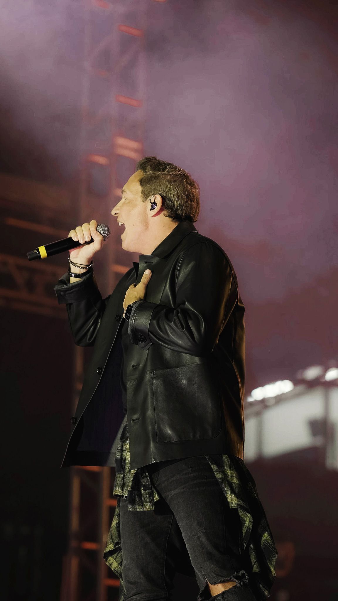 Um artista vestindo uma jaqueta de couro preta canta em um microfone em um palco com iluminação difusa.