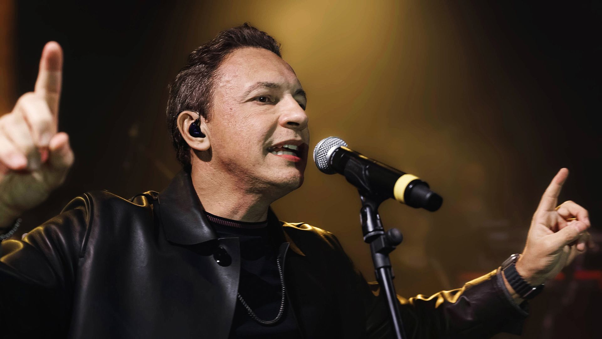 Um cantor se apresentando no palco, segurando um microfone e gesticulando apontadas para cima em um ambiente com pouca luz.