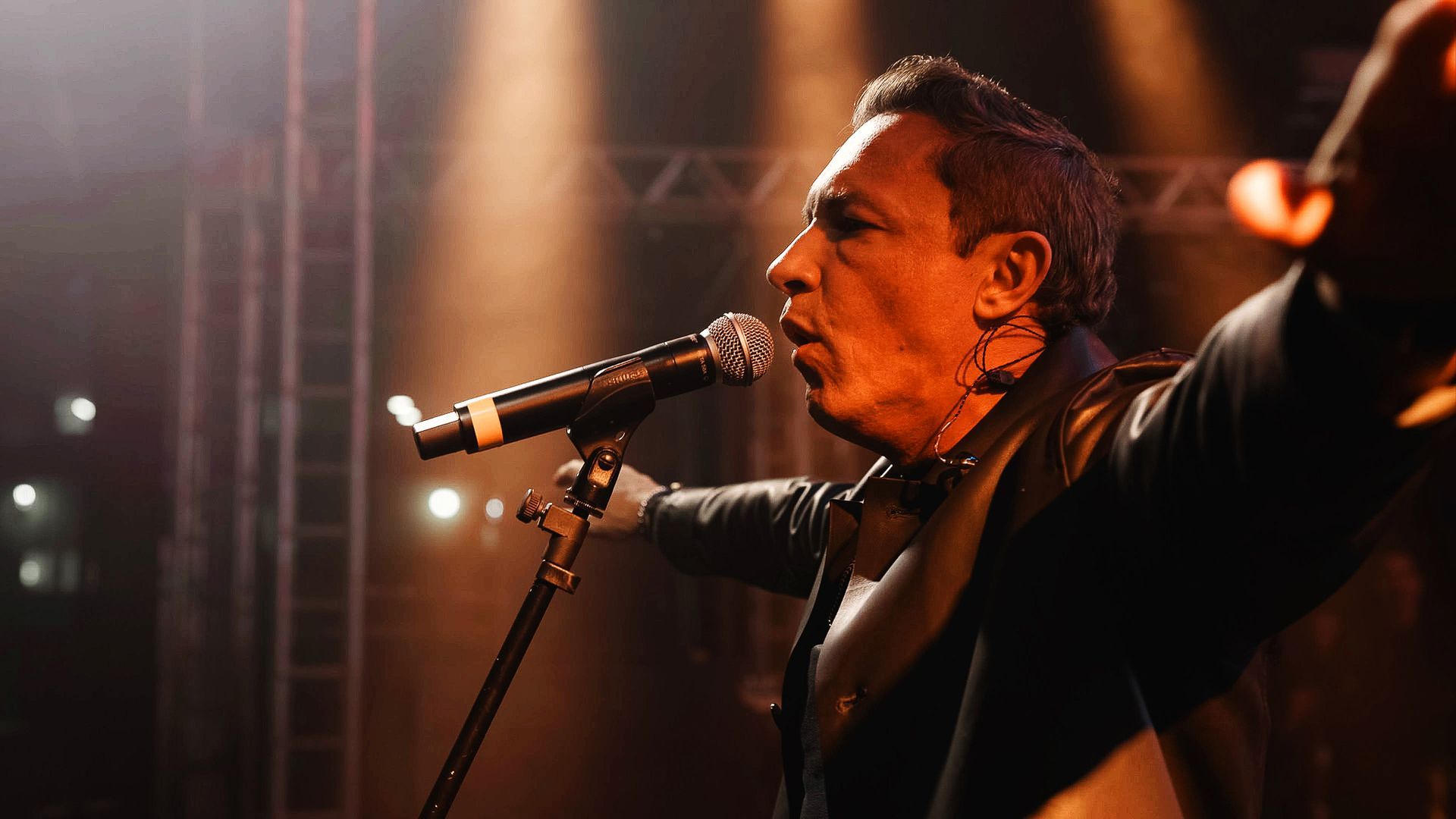 Um músico cantando ao microfone no palco, com os braços estendidos sob holofotes aconchegantes.