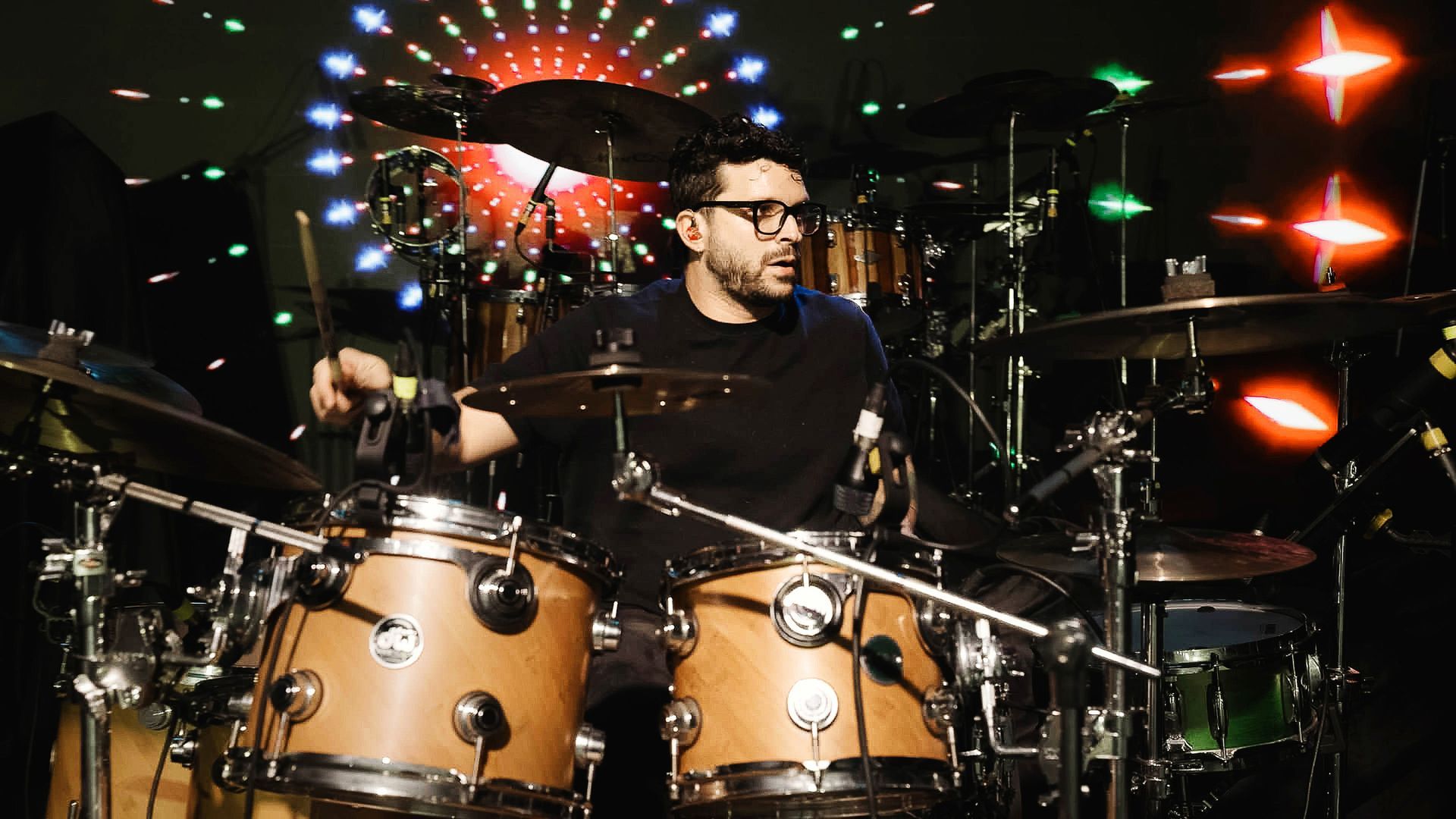 Um baterista de óculos toca bateria em um palco com luzes circulares brilhantes e coloridas ao fundo.