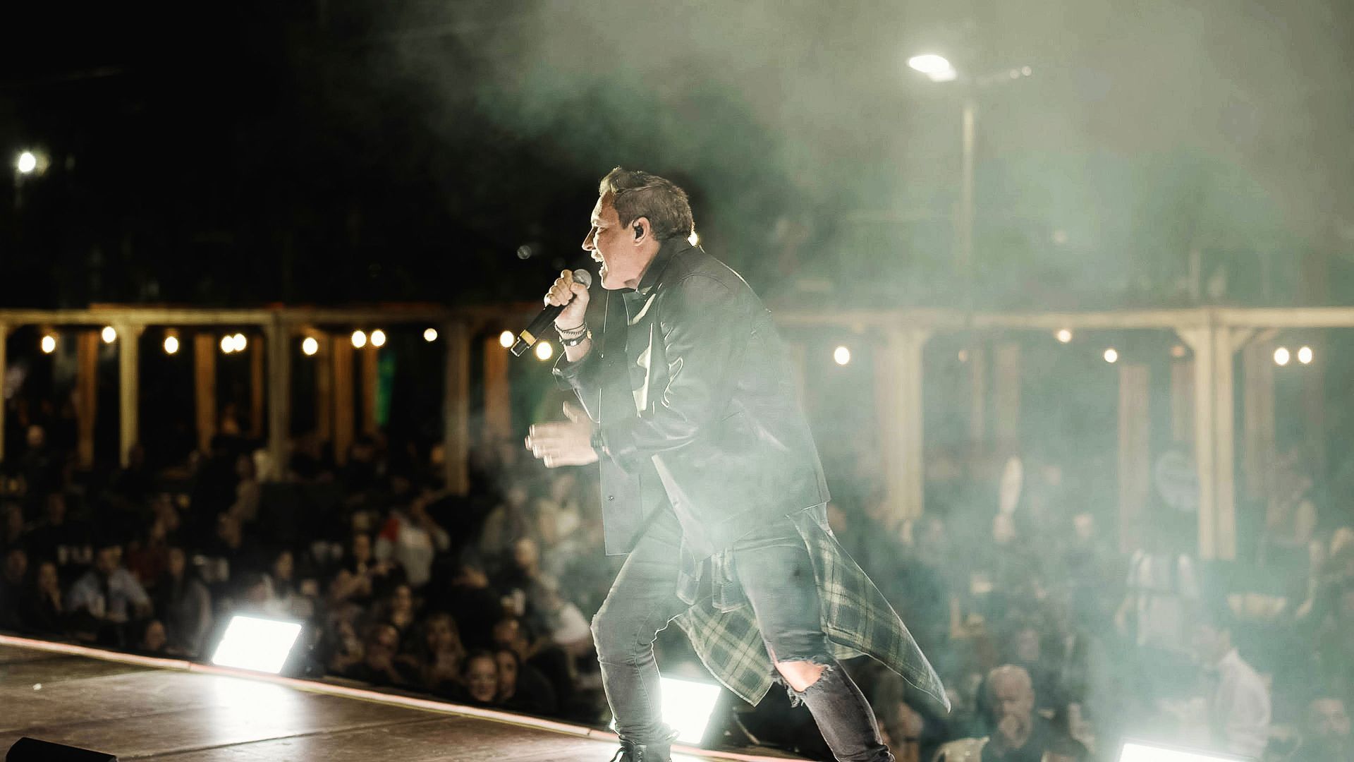 Um artista de jaqueta de couro está em um palco segurando um microfone, sua silhueta recortada contra as luzes do palco.