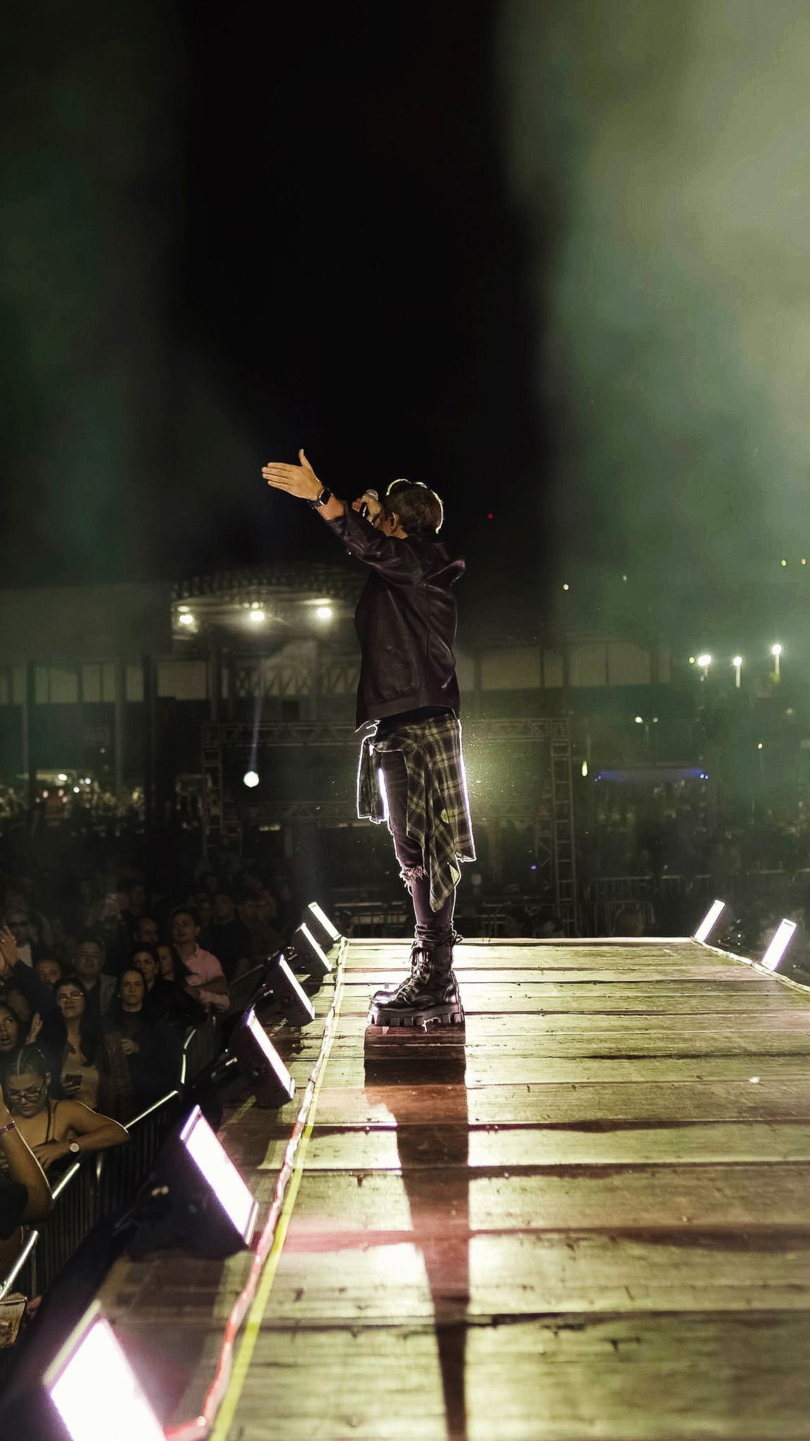 Um artista está em um palco com os braços estendidos em direção à plateia durante um show noturno ao ar livre.