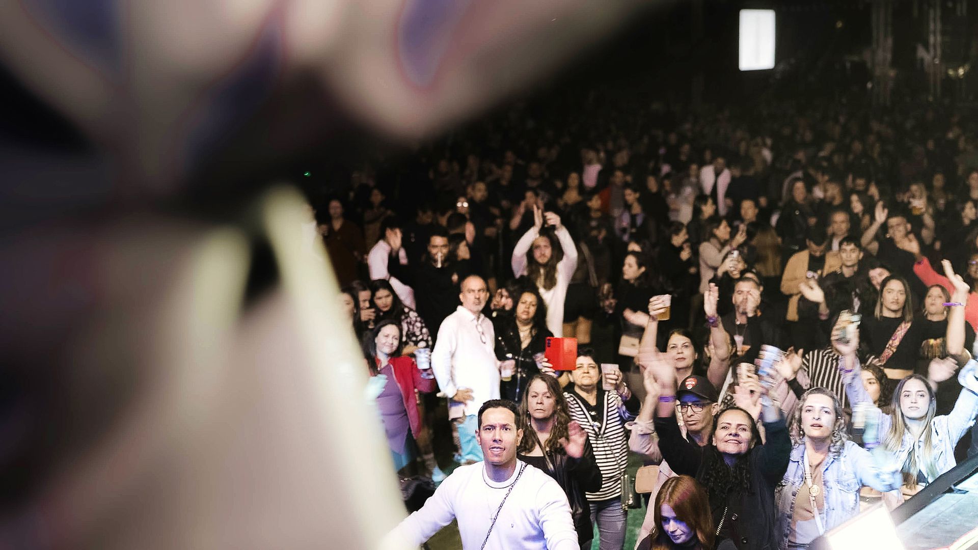 Uma multidão de pessoas em um concerto noturno, com alguns membros da plateia aplaudindo e levantando as mãos.