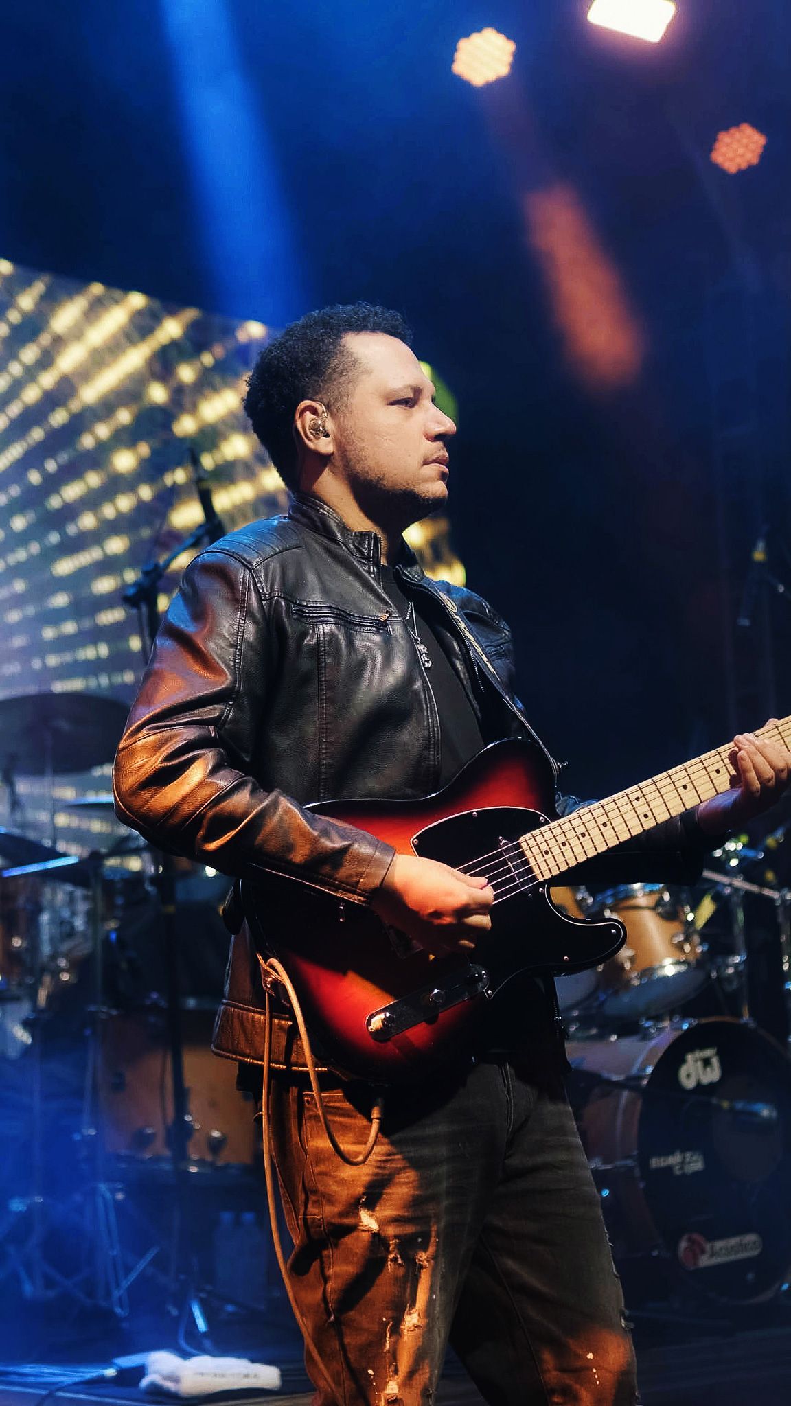 Um músico de jaqueta de couro toca uma guitarra elétrica sunburst em um palco com luzes de concerto e bateria.