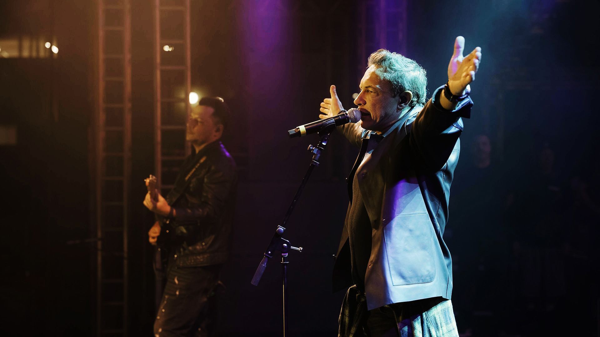 Um cantor com os braços erguidos se apresenta no palco com um guitarrista ao seu lado, sob luzes de concerto azuis e roxas.