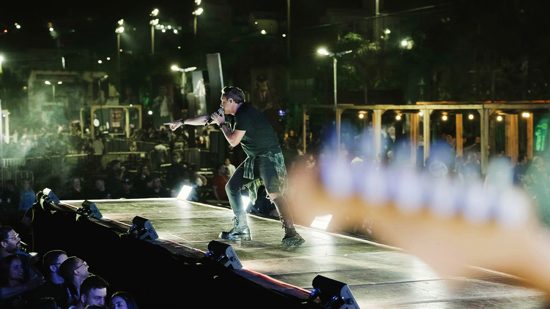 Um artista canta em um microfone em um palco ao ar livre à noite, apontando para uma multidão sob luzes de palco brilhantes.