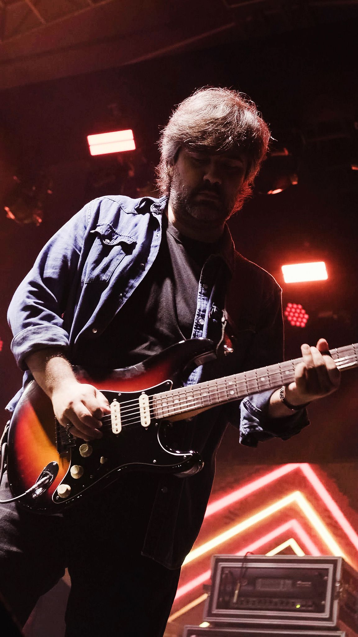 Um guitarrista toca uma guitarra elétrica sunburst em um palco com estruturas de luzes neon ao fundo.