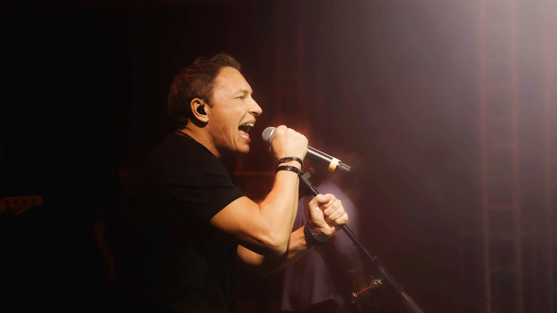 Um artista vestindo uma camisa preta canta em um microfone em um palco pouco iluminado, com um holofote brilhante ao fundo.