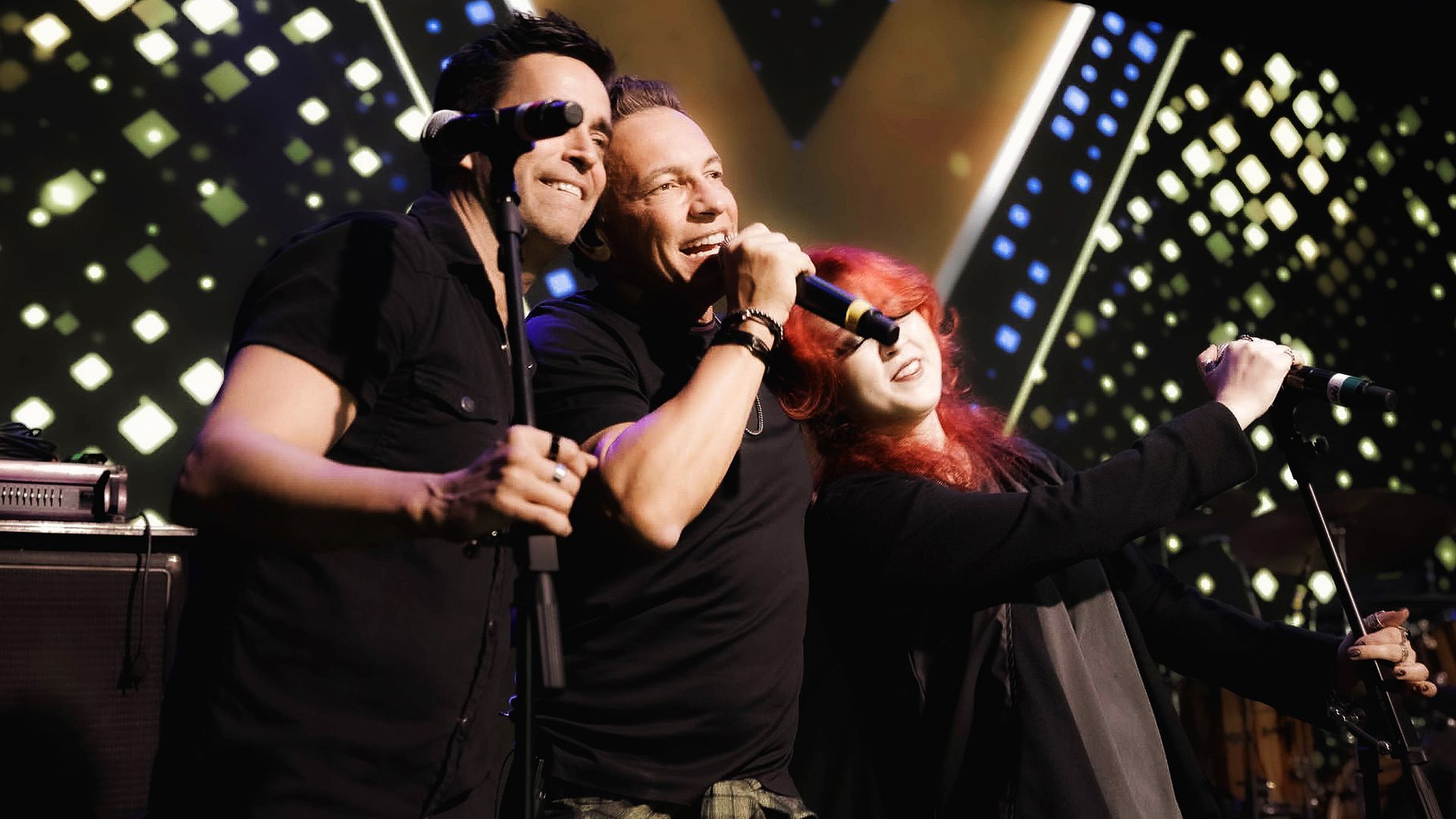 Três músicos se apresentam no palco sob luzes brilhantes, cantando em microfones com poses expressivas e enérgicas.