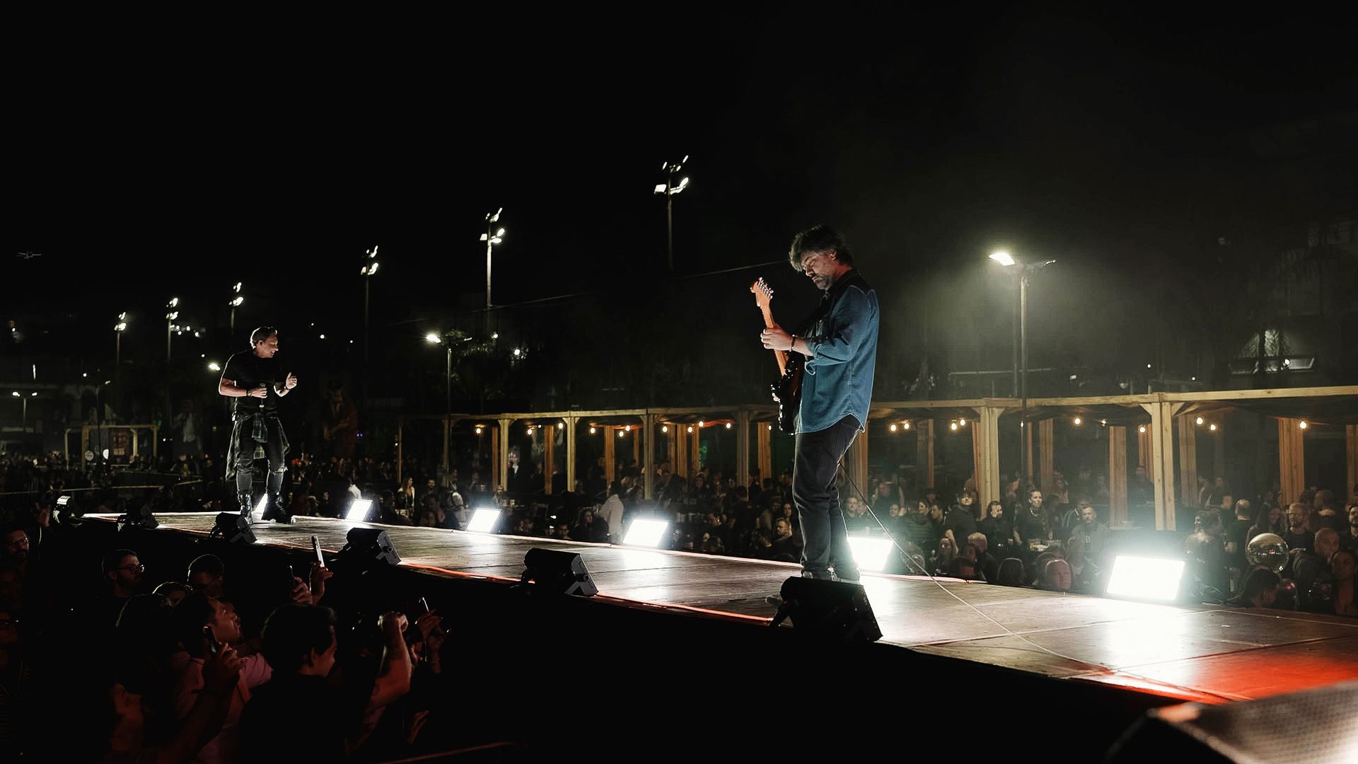 Dois músicos se apresentam em um palco externo bem iluminado à noite, diante de uma grande plateia no escuro.