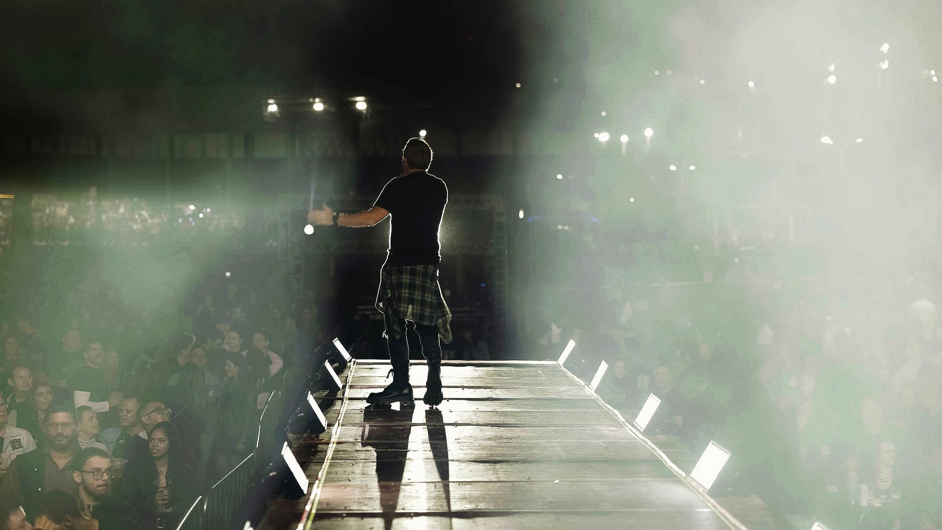 Um artista está em pé na passarela do palco, de frente para a plateia, sob luzes dramáticas e névoa.