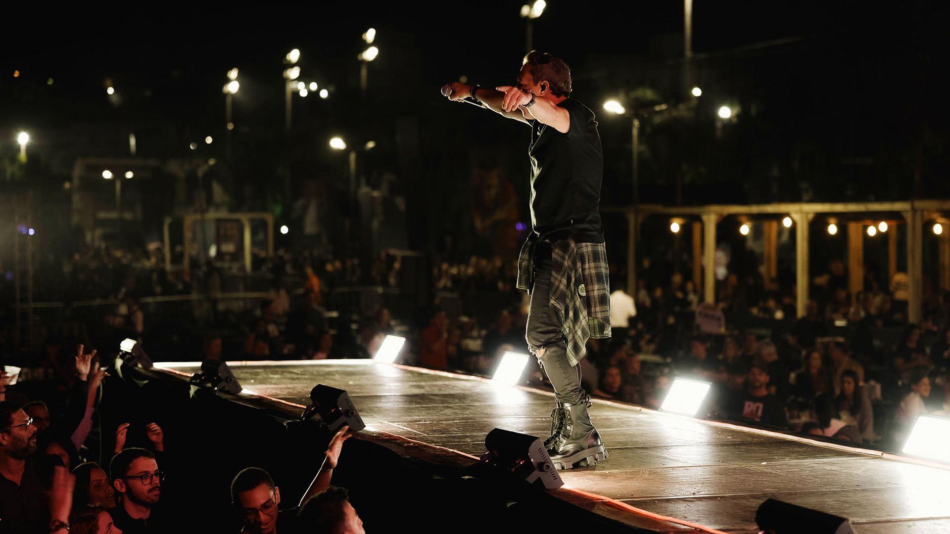 Um artista se apresenta em um palco bem iluminado à noite, com os braços estendidos, de frente para a plateia em um evento.