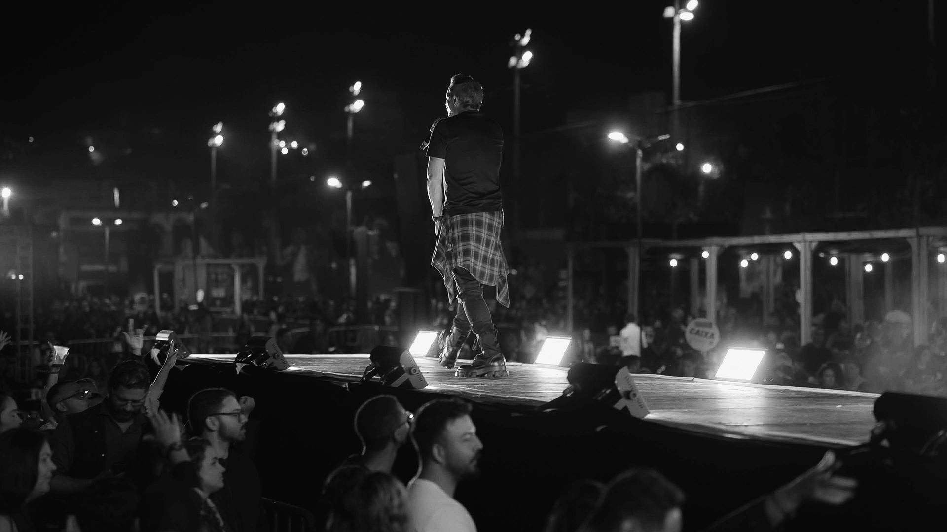 Uma artista se apresenta em um palco, iluminada por luzes, de frente para uma plateia, em uma foto em preto e branco.