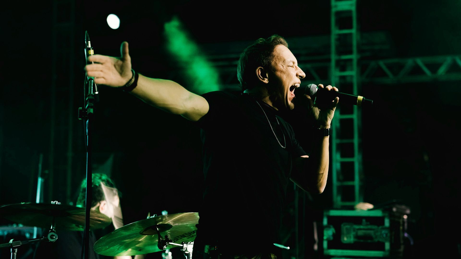 Um músico com os braços estendidos canta em um microfone em um palco pouco iluminado por holofotes verdes.
