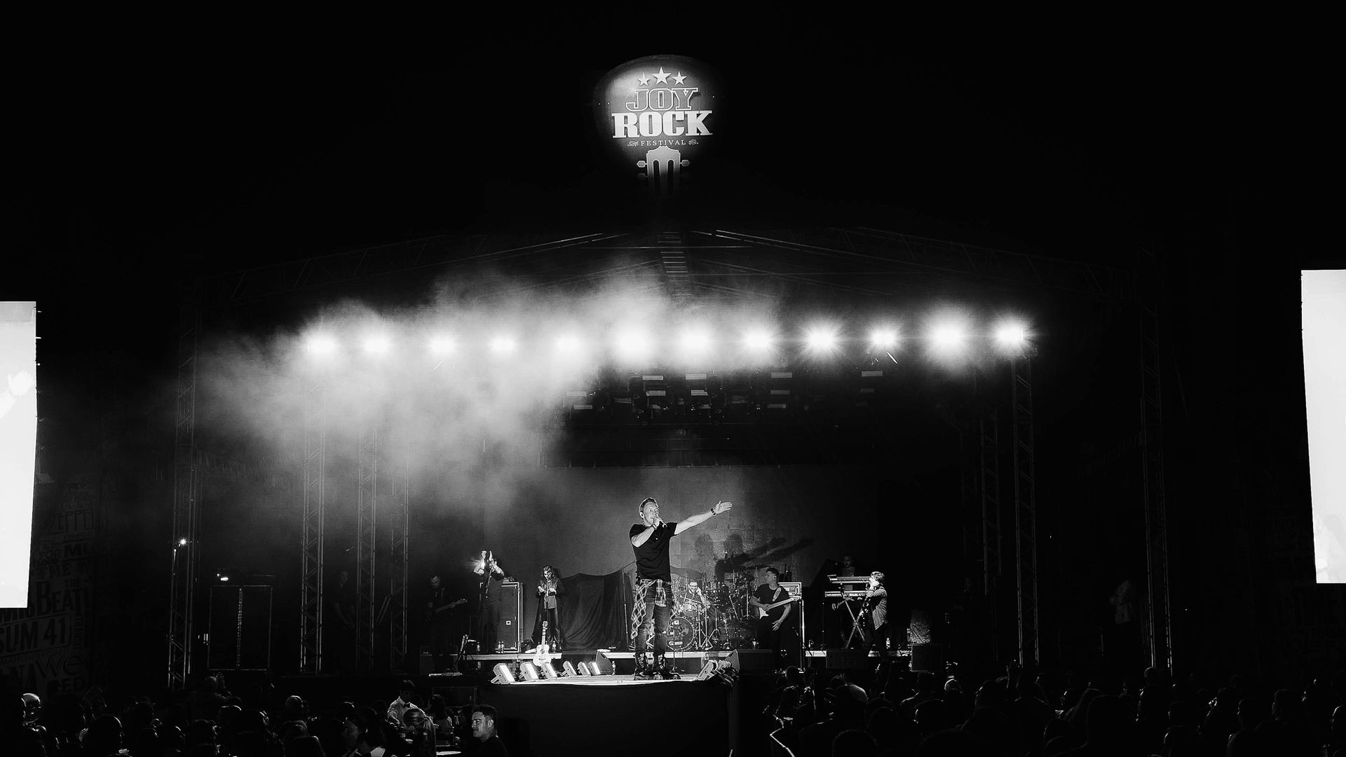 Um artista no palco de um show estende a mão para a multidão sob holofotes brilhantes e um grande logotipo do festival
