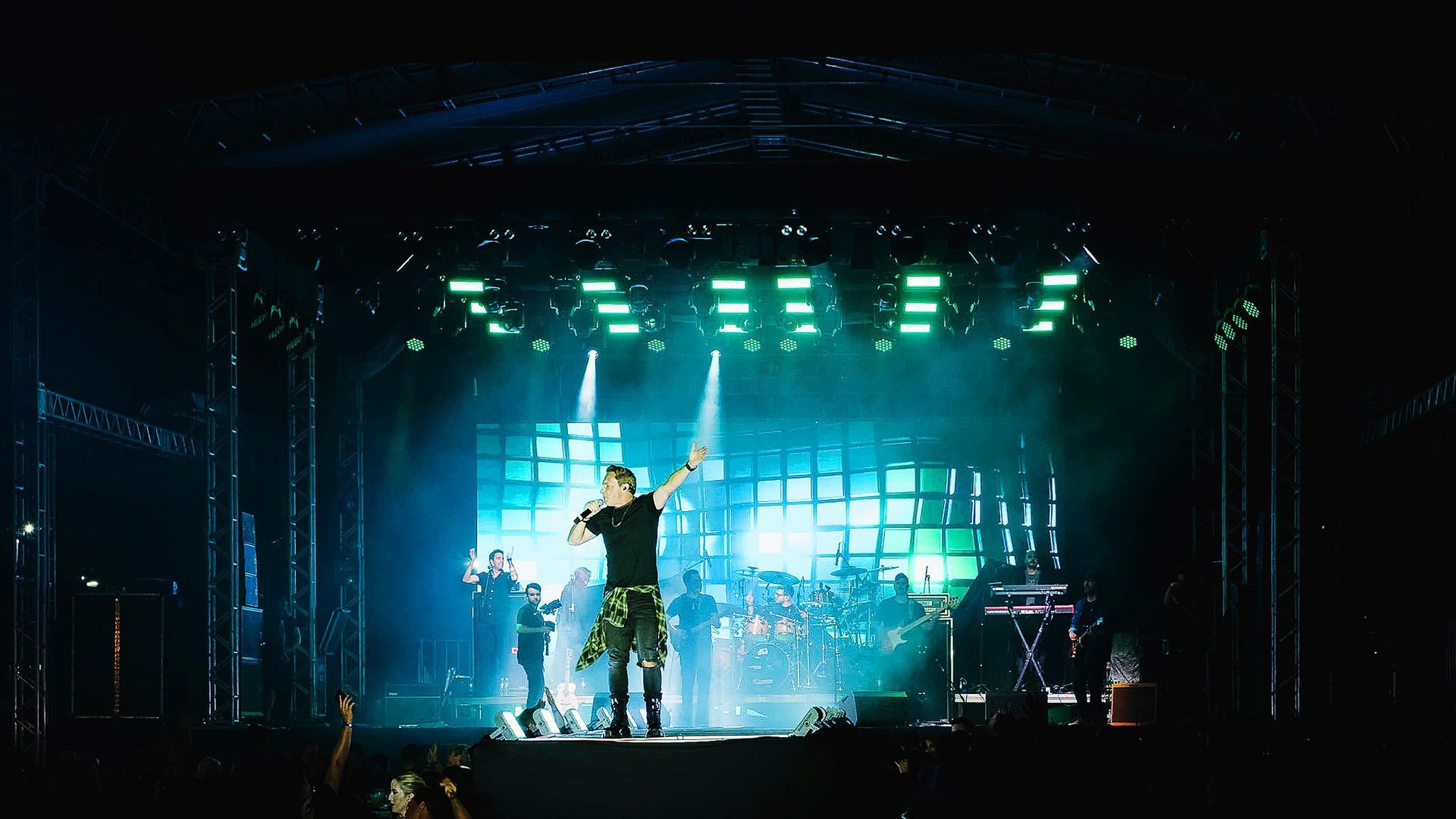 O vocalista principal se apresenta em um palco iluminado por holofotes, com uma banda e uma tela digital ao fundo.