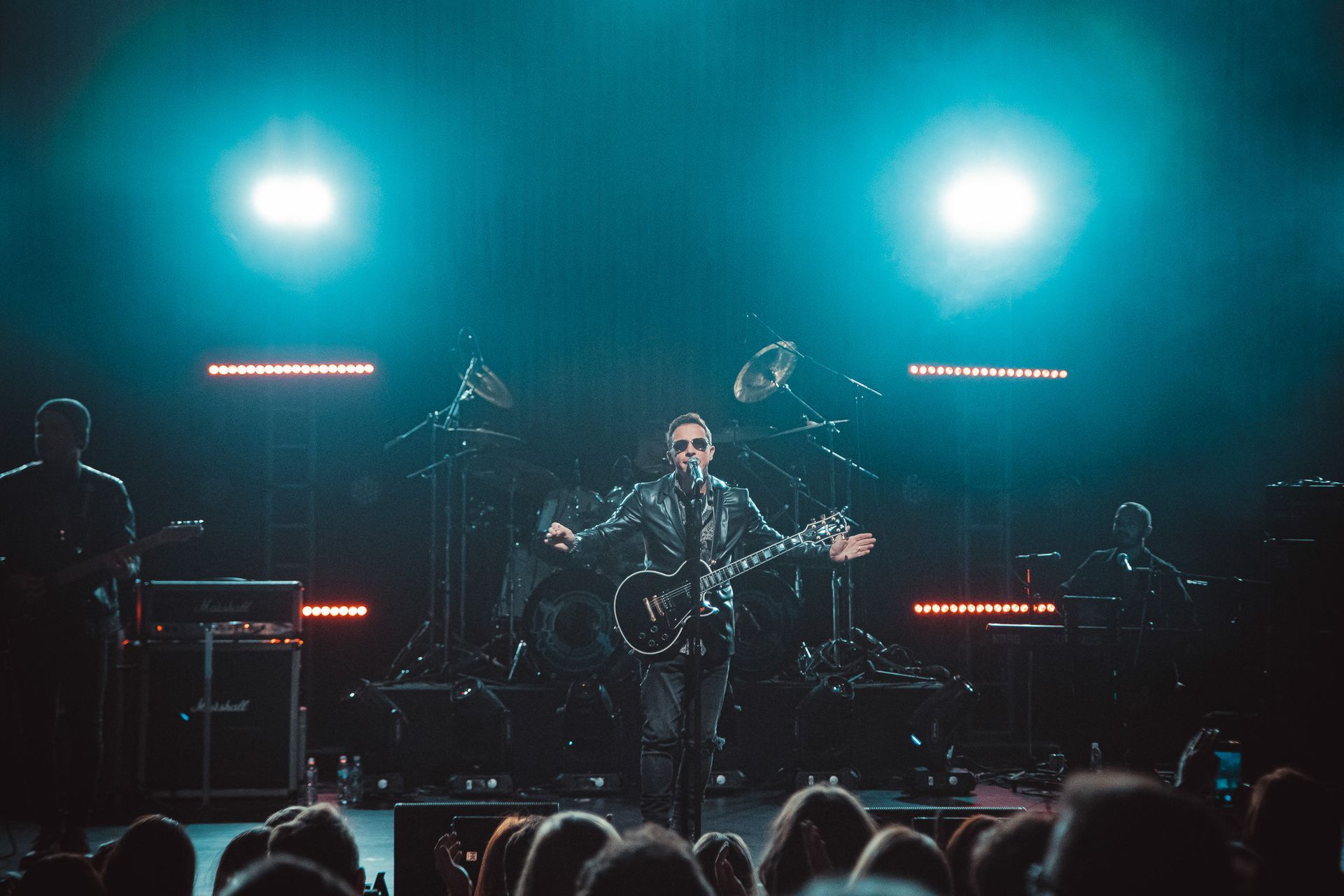 O vocalista, vestindo jaqueta de couro, toca guitarra no palco sob holofotes, apresentando-se para uma plateia.
