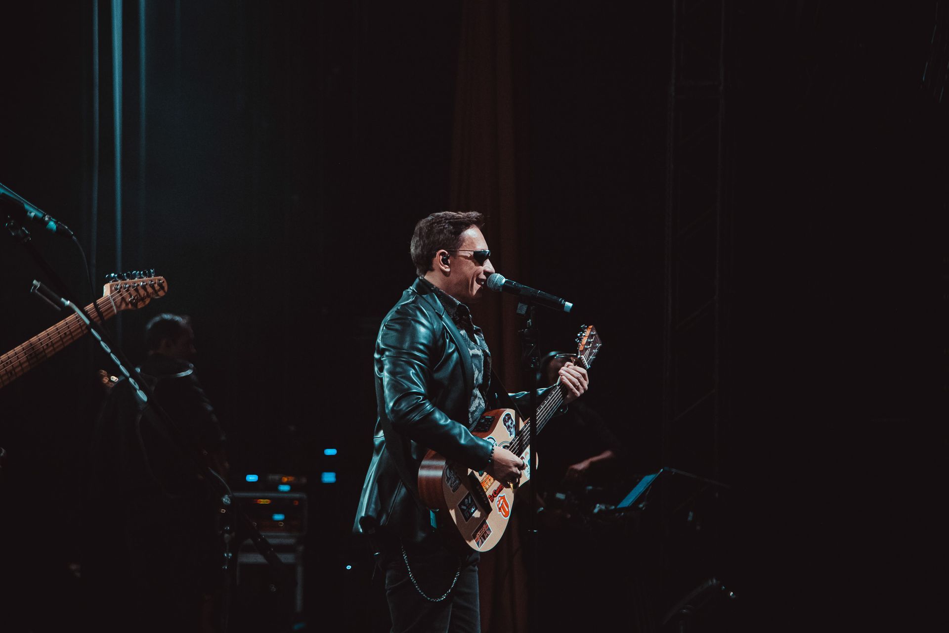 Um artista vestindo jaqueta de couro e óculos escuros toca violão enquanto canta ao microfone em um palco.