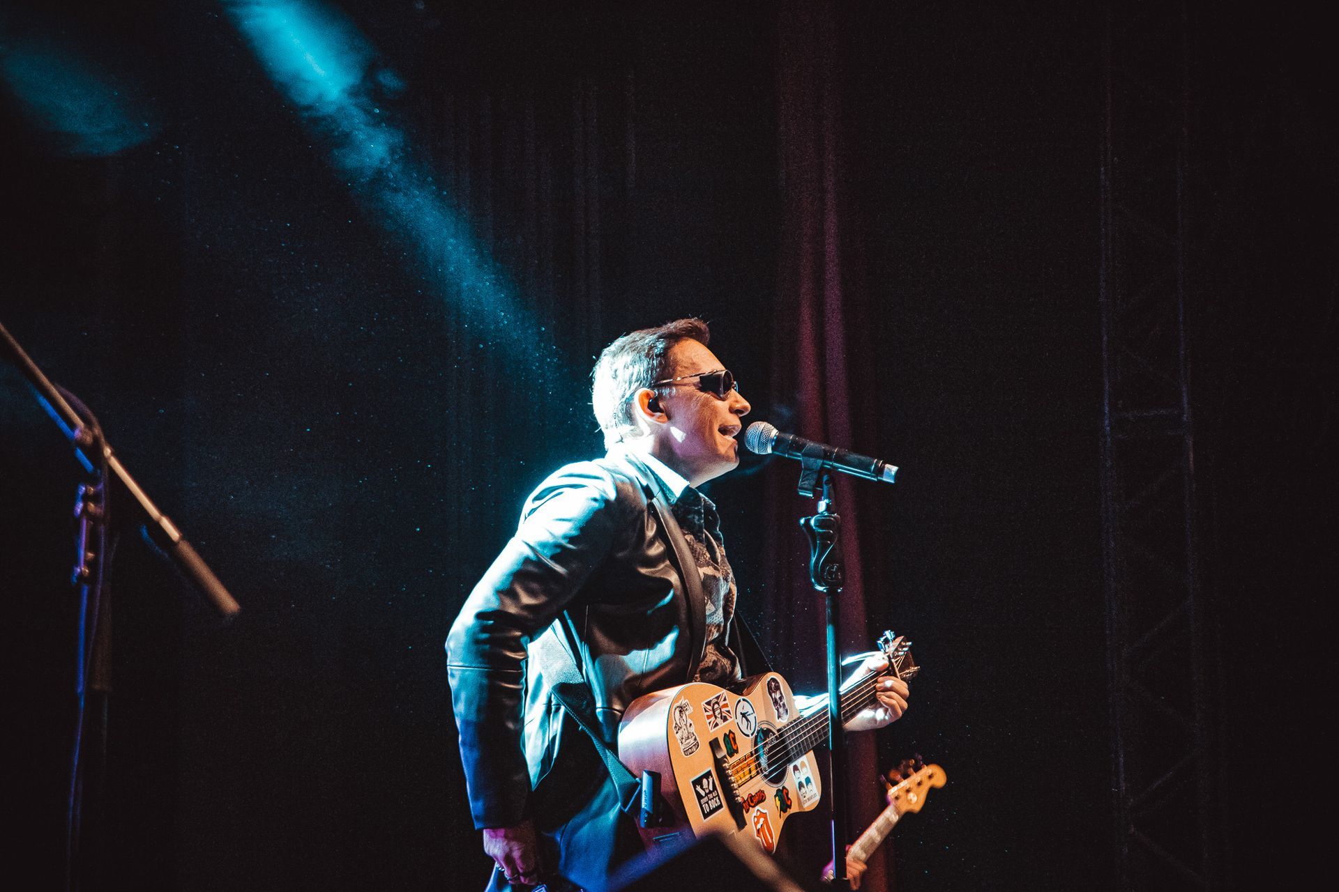 Um músico de óculos escuros se apresenta em um palco escuro, tocando uma guitarra coberta de adesivos em um microfone.