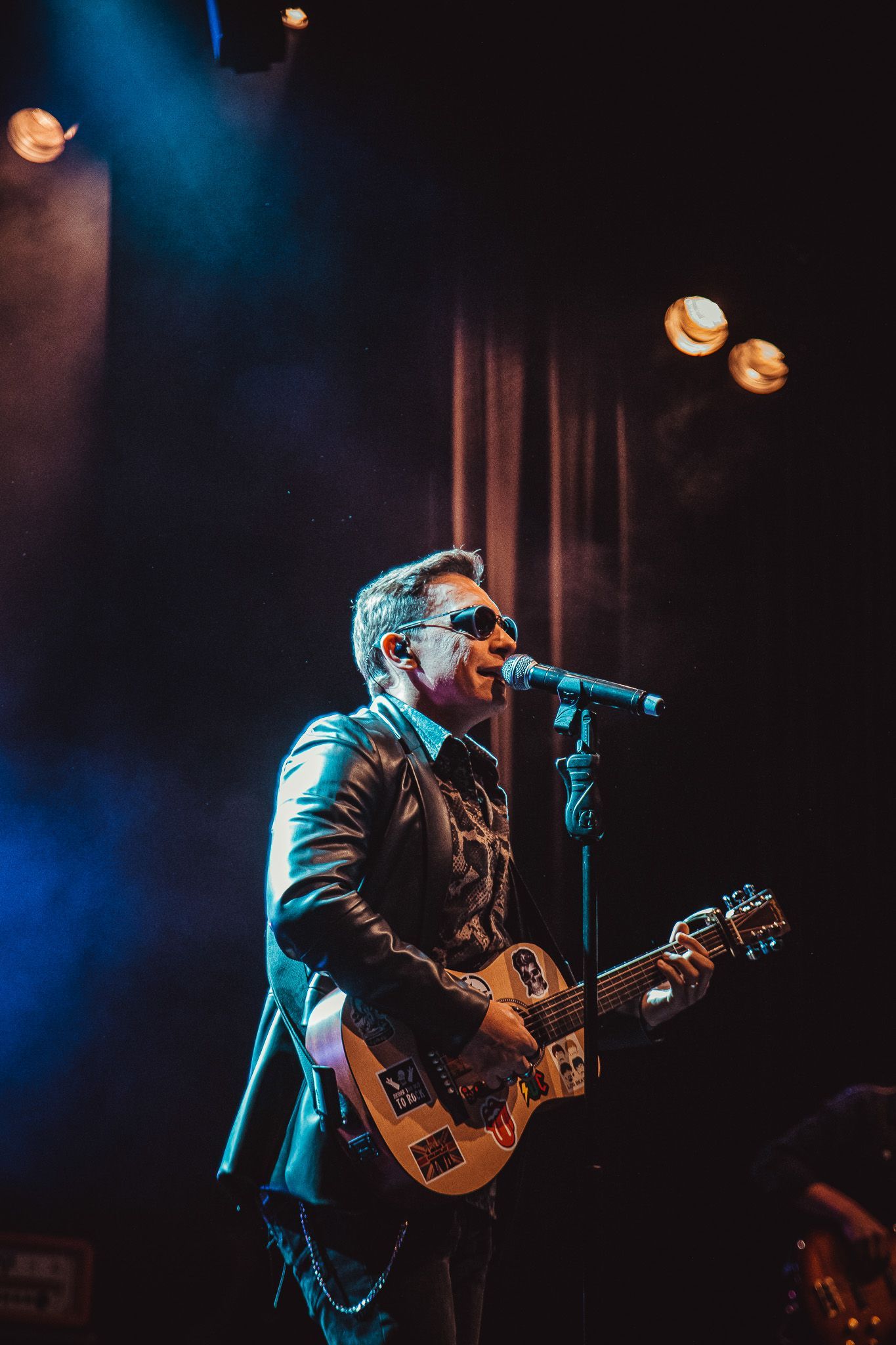 Um músico de jaqueta de couro se apresenta no palco com guitarra e microfone sob iluminação cênica azul e quente.