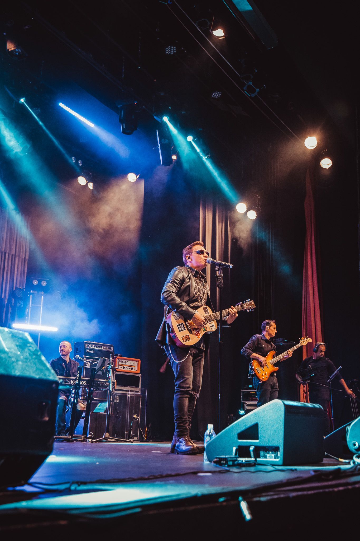 Uma banda se apresenta em um palco sob holofotes azuis e brancos brilhantes, com músicos tocando guitarras e cantando.