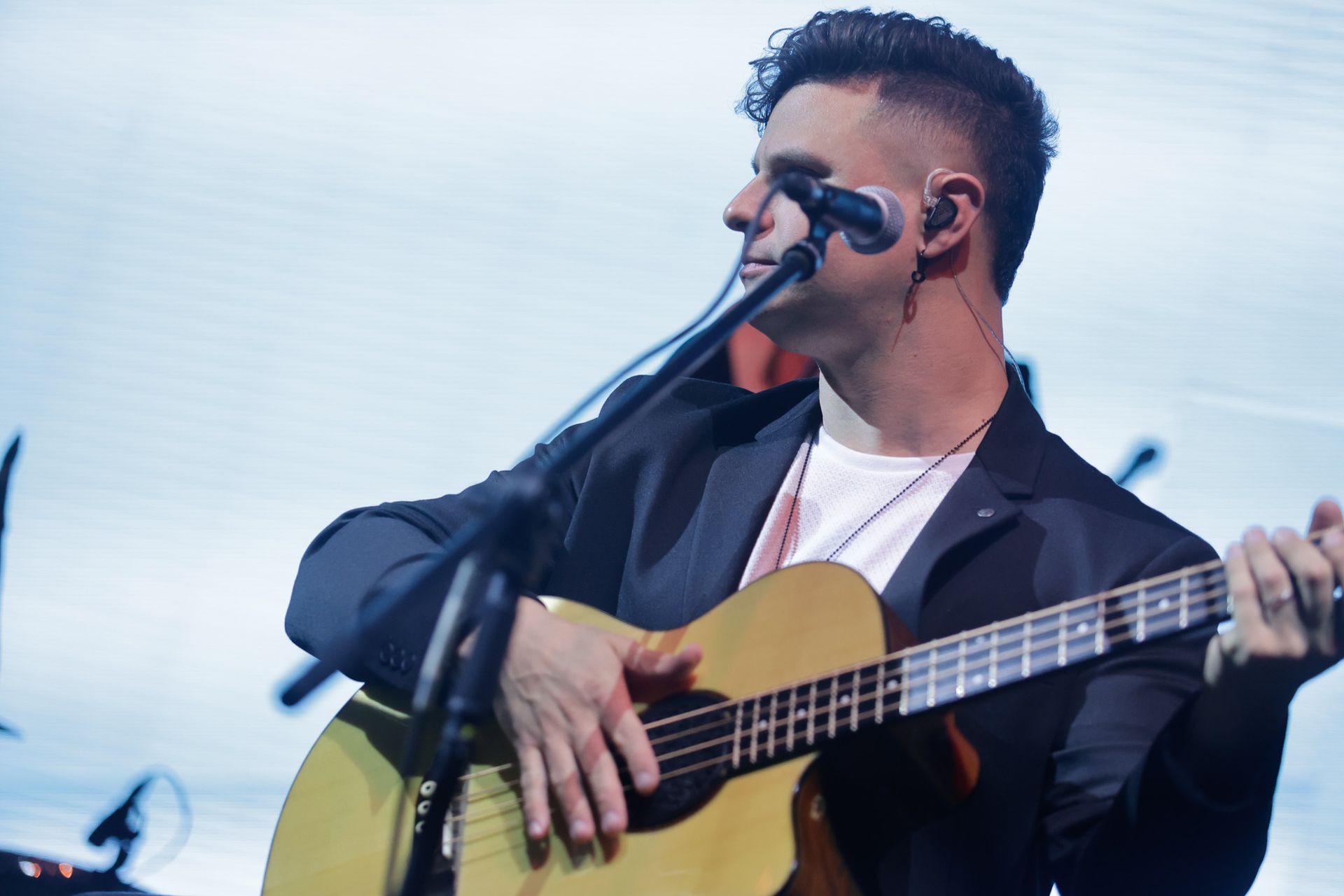 Um artista tocando violão no palco, vestindo um blazer e com um microfone.