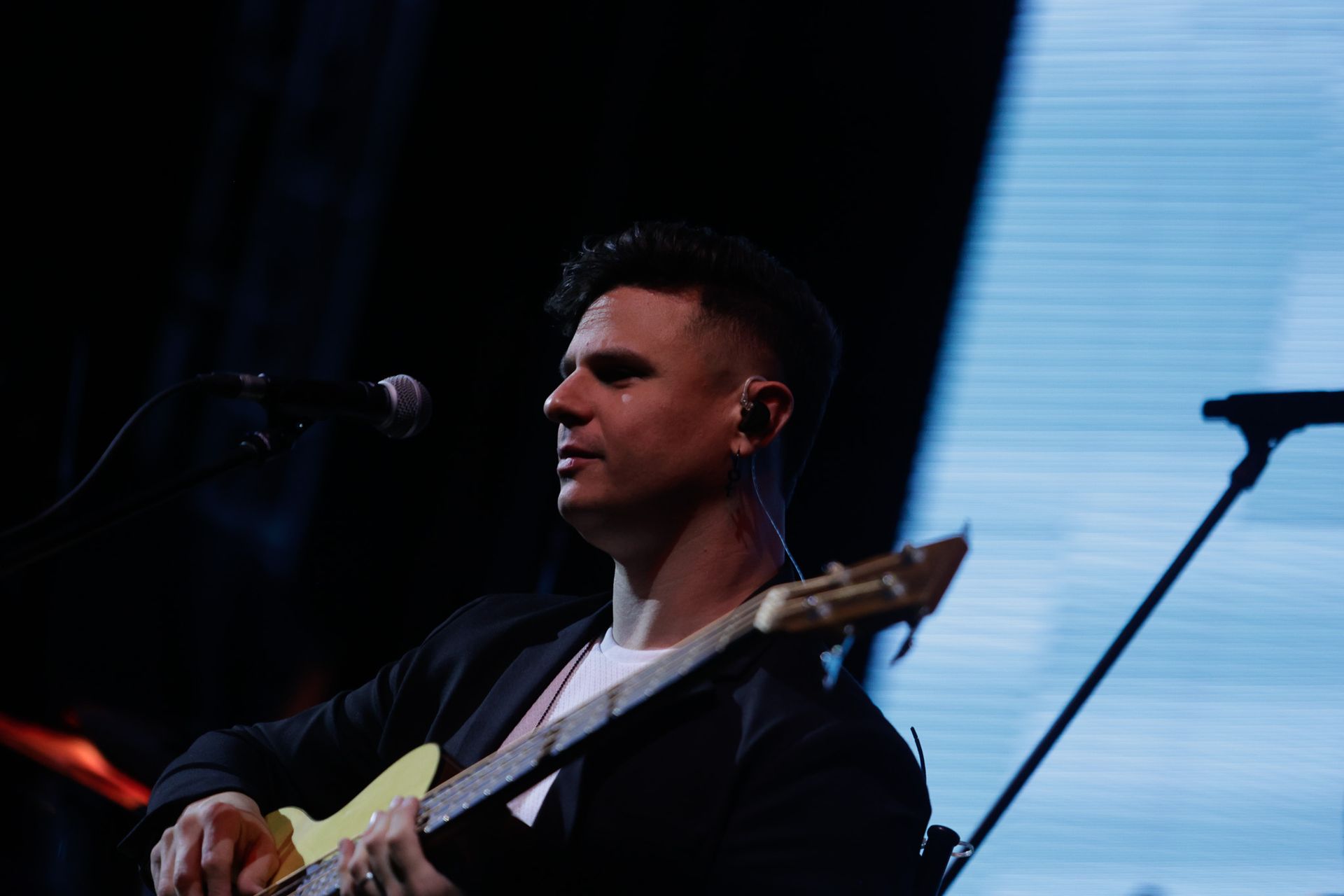 Um artista tocando baixo e cantando em um show, com luzes de palco ao fundo.