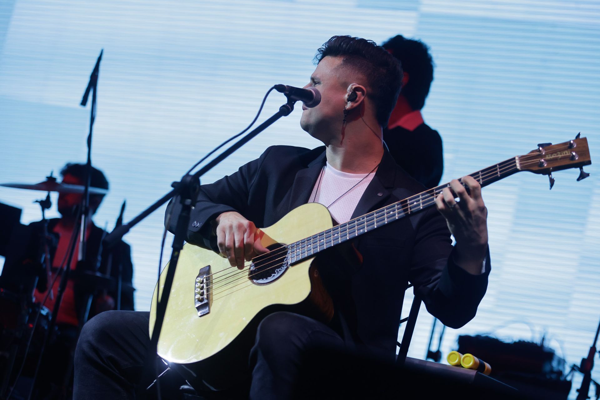Um artista tocando baixo e cantando ao microfone no palco; luzes brilhantes e banda ao fundo.