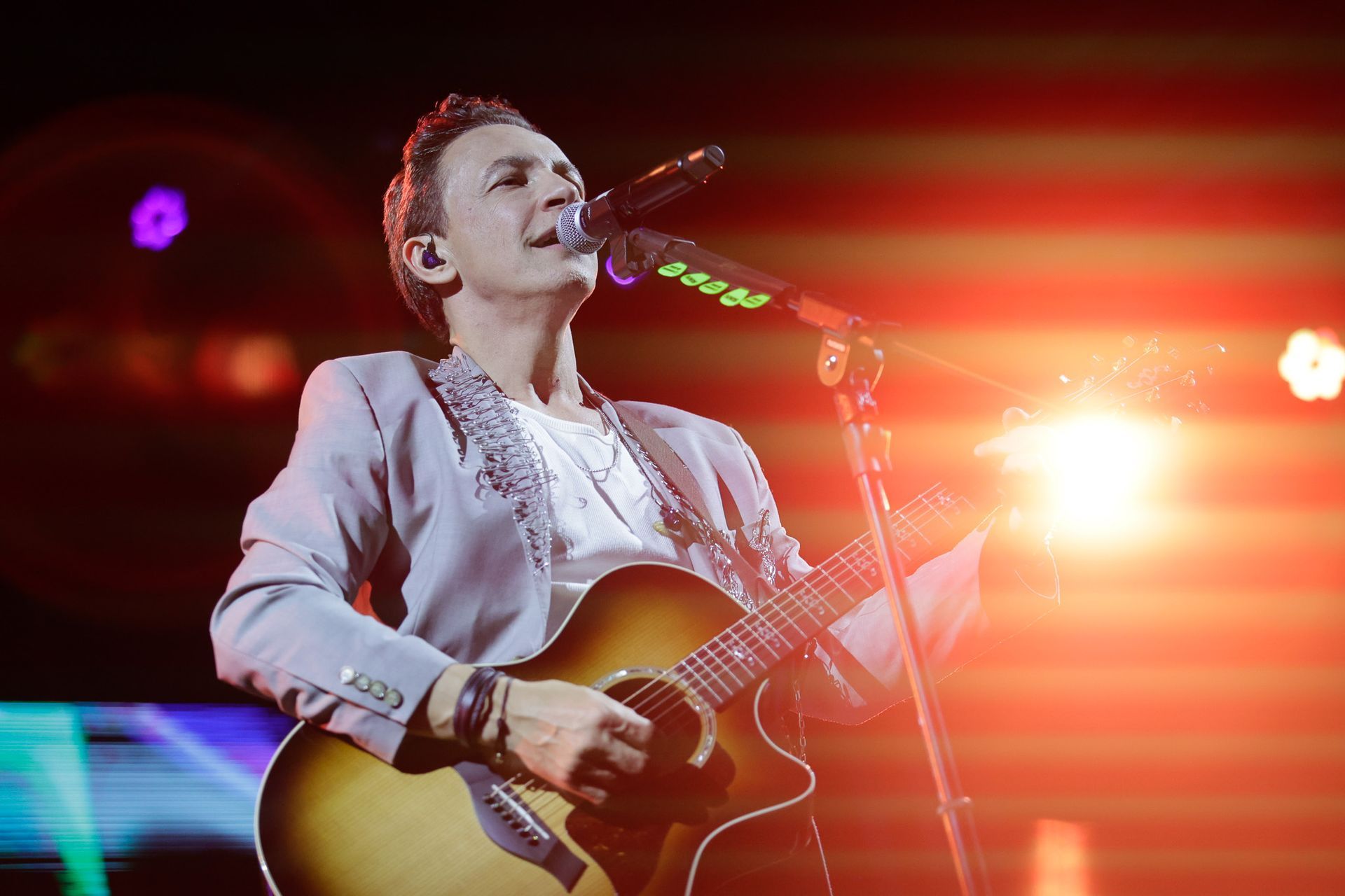 Um artista tocando violão e cantando ao microfone no palco sob luzes fortes.