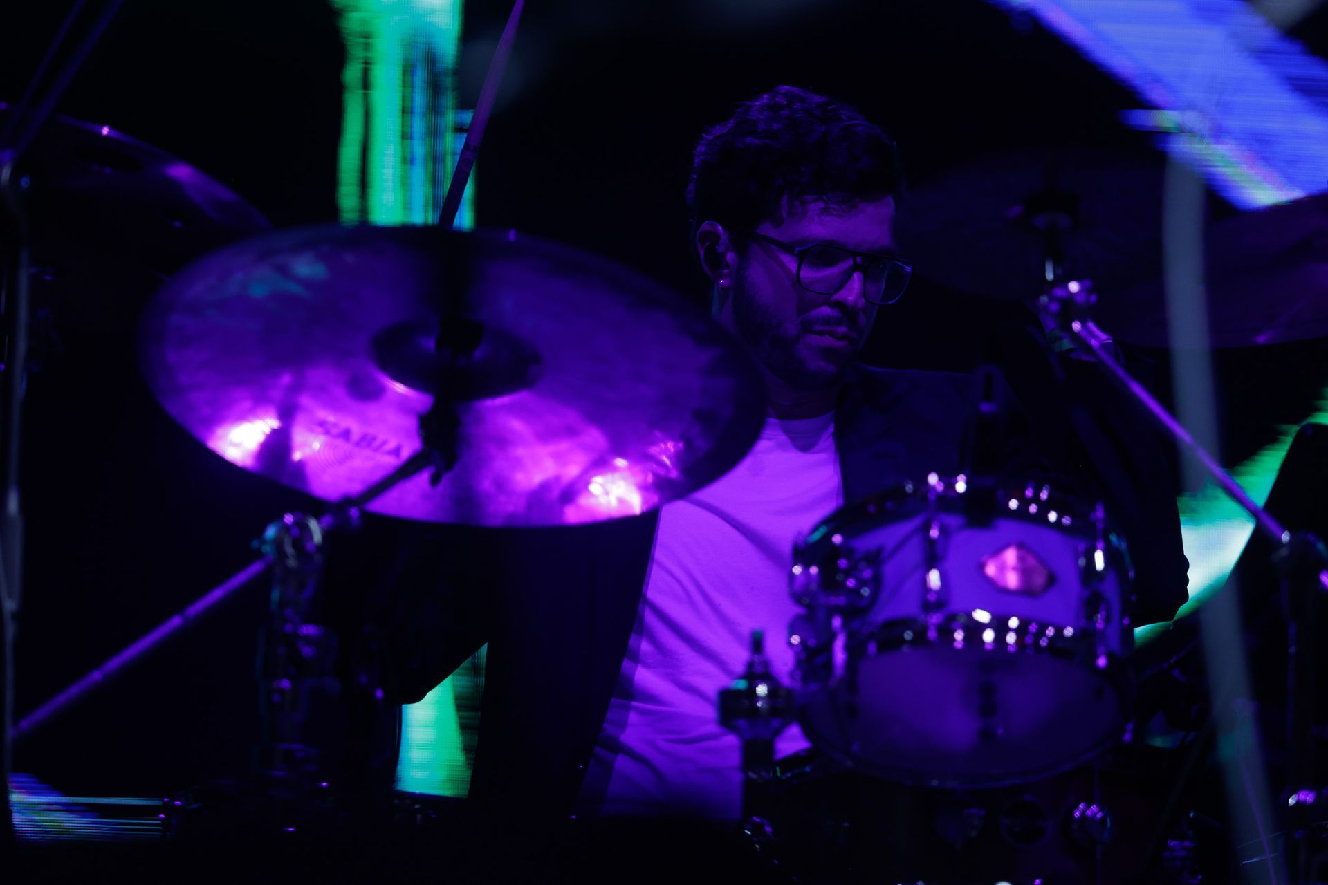 Baterista tocando bateria no palco, iluminado com luzes roxas e verdes.